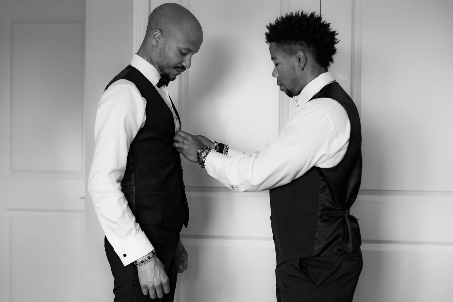 Two people in matching black vests helping each other prepare formal attire.