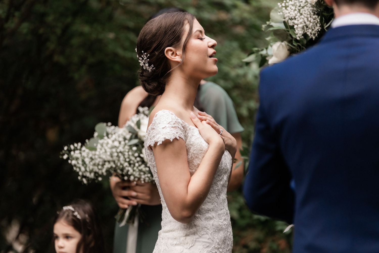 Intimate garden wedding ceremony moment with bride in lace gown holding white floral bouquet.