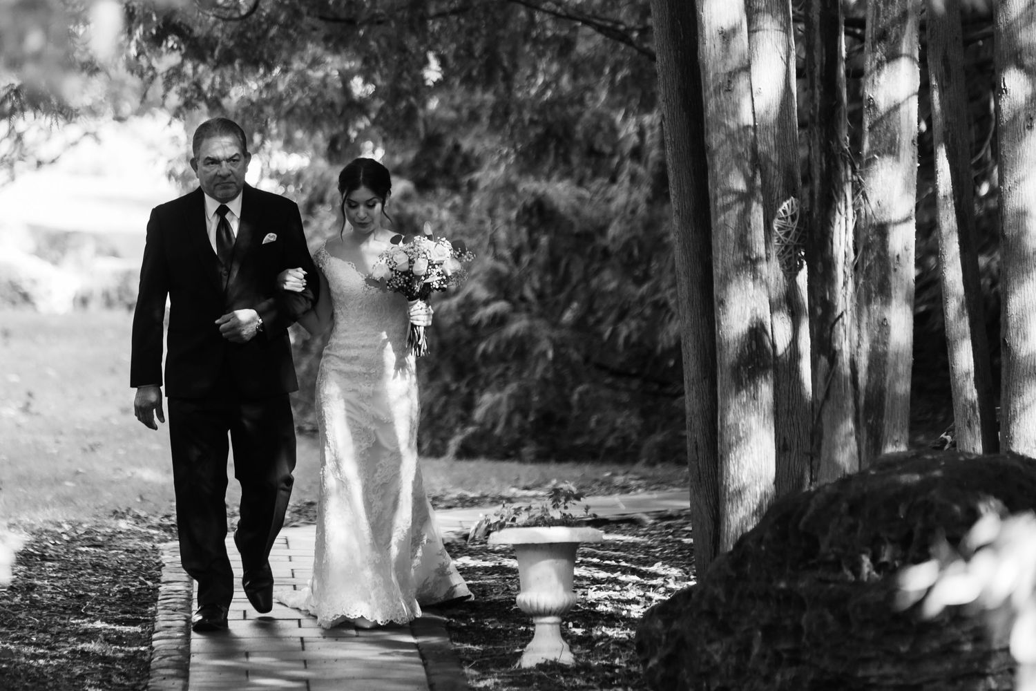 A black and white image shows a bride walking down a wet garden path with her escort.