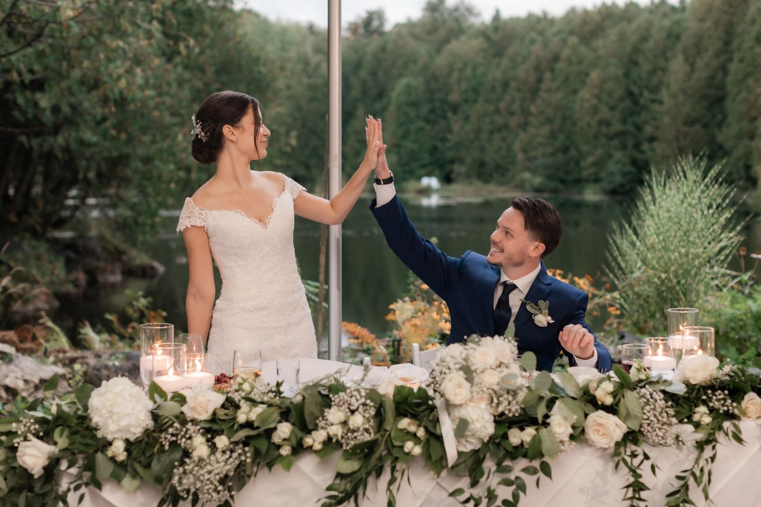 Newlyweds give each other a high-five at their wedding reception behind a floral-decorated table.