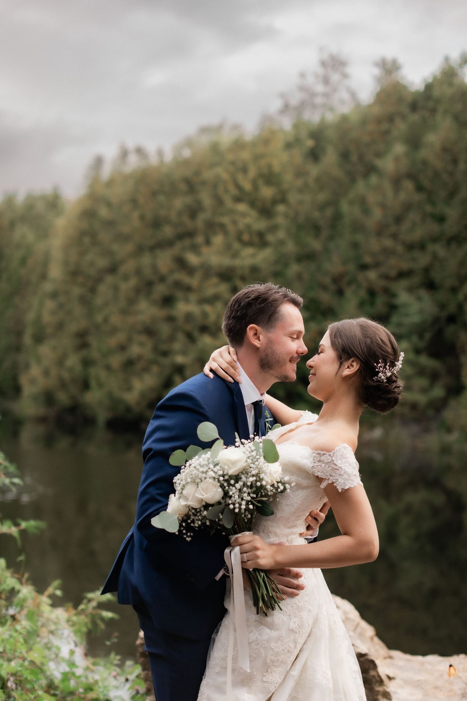 A romantic wedding moment captured by a lake with floral bouquet and autumn trees in background.