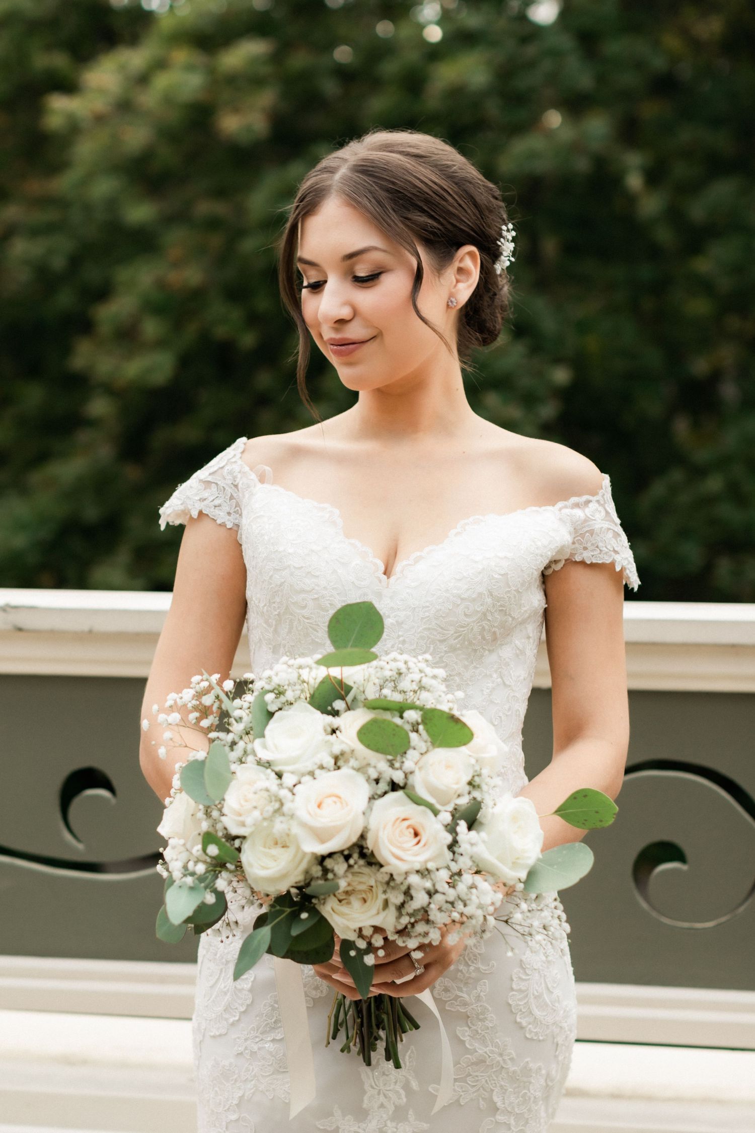 Bride in lace off-shoulder wedding dress holding elegant white rose and eucalyptus bouquet on outdoor balcony.