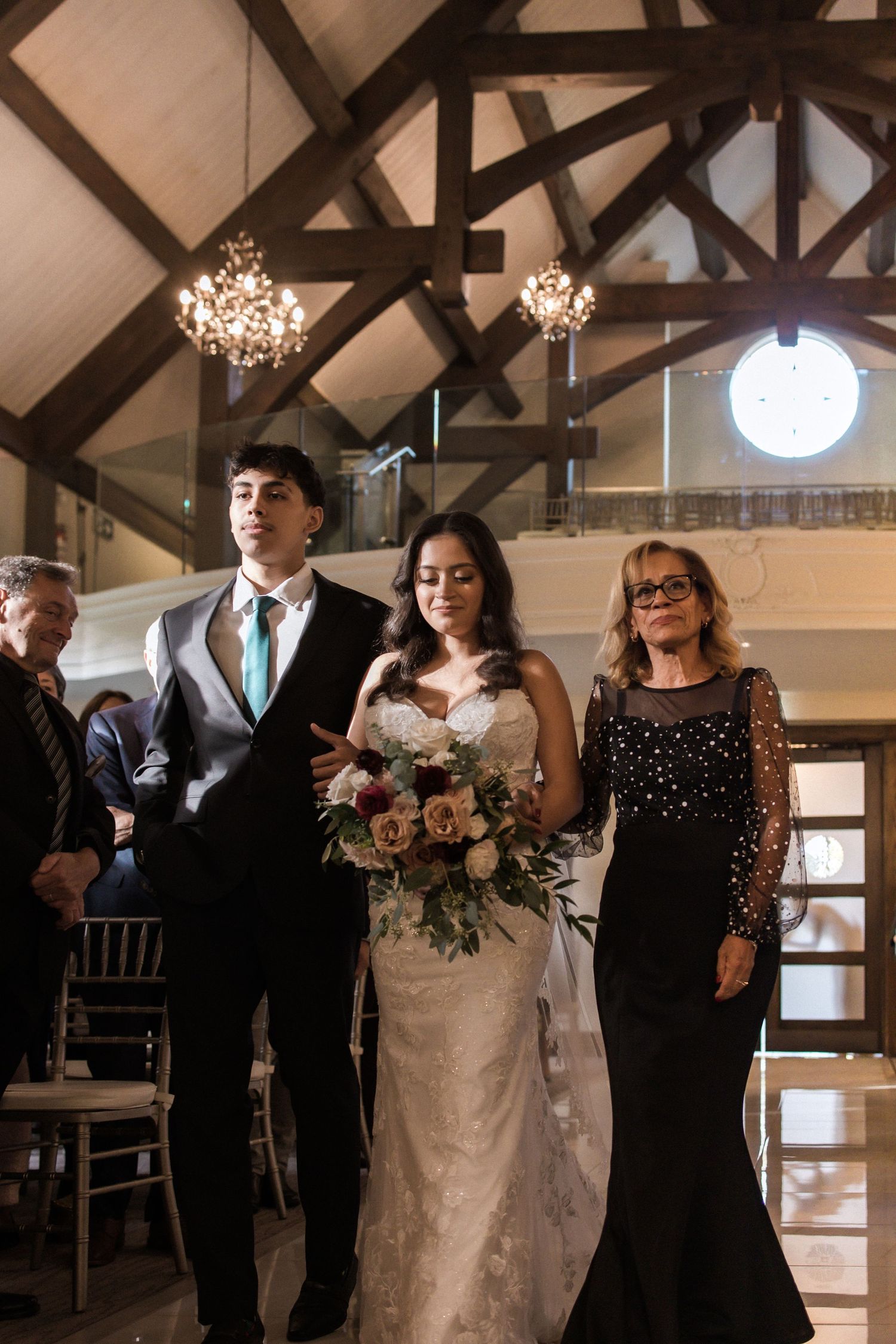 Wedding party walks down the aisle in a chapel featuring exposed beams and crystal chandeliers.