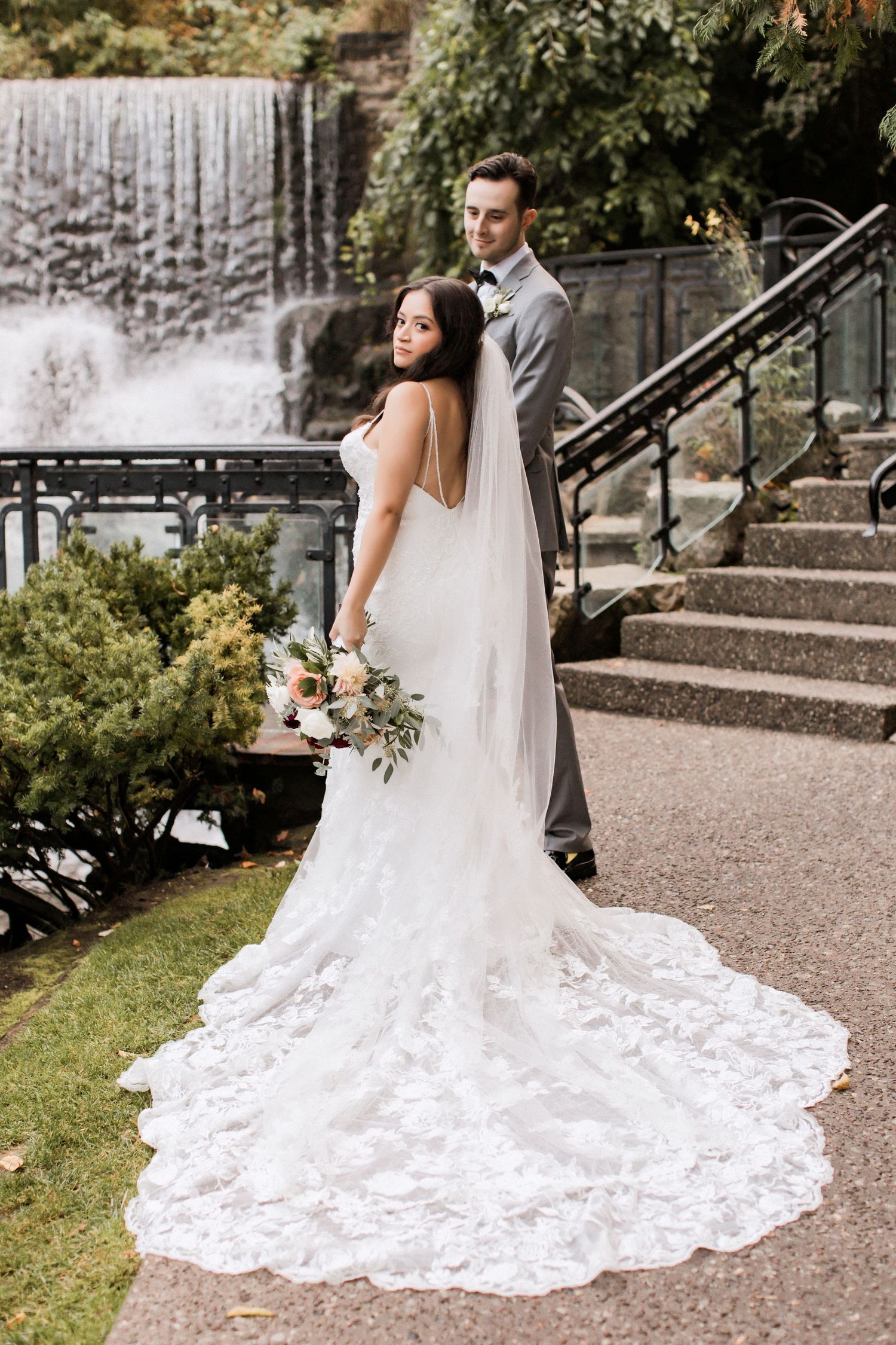 Elegant bride and groom pose by cascading waterfall with flowing lace wedding dress train.