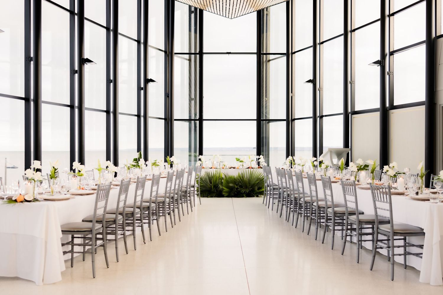 Elegant wedding reception layout with two long tables and silver chairs in bright modern venue with floor-to-ceiling windows.