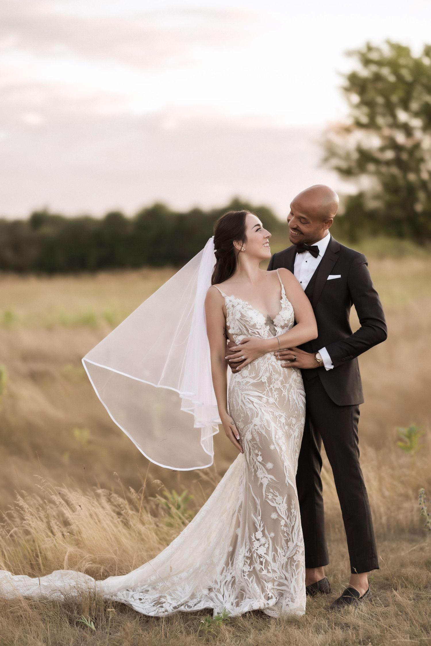 Elegant wedding couple poses in a meadow with bride's detailed lace dress and flowing veil catching the light.