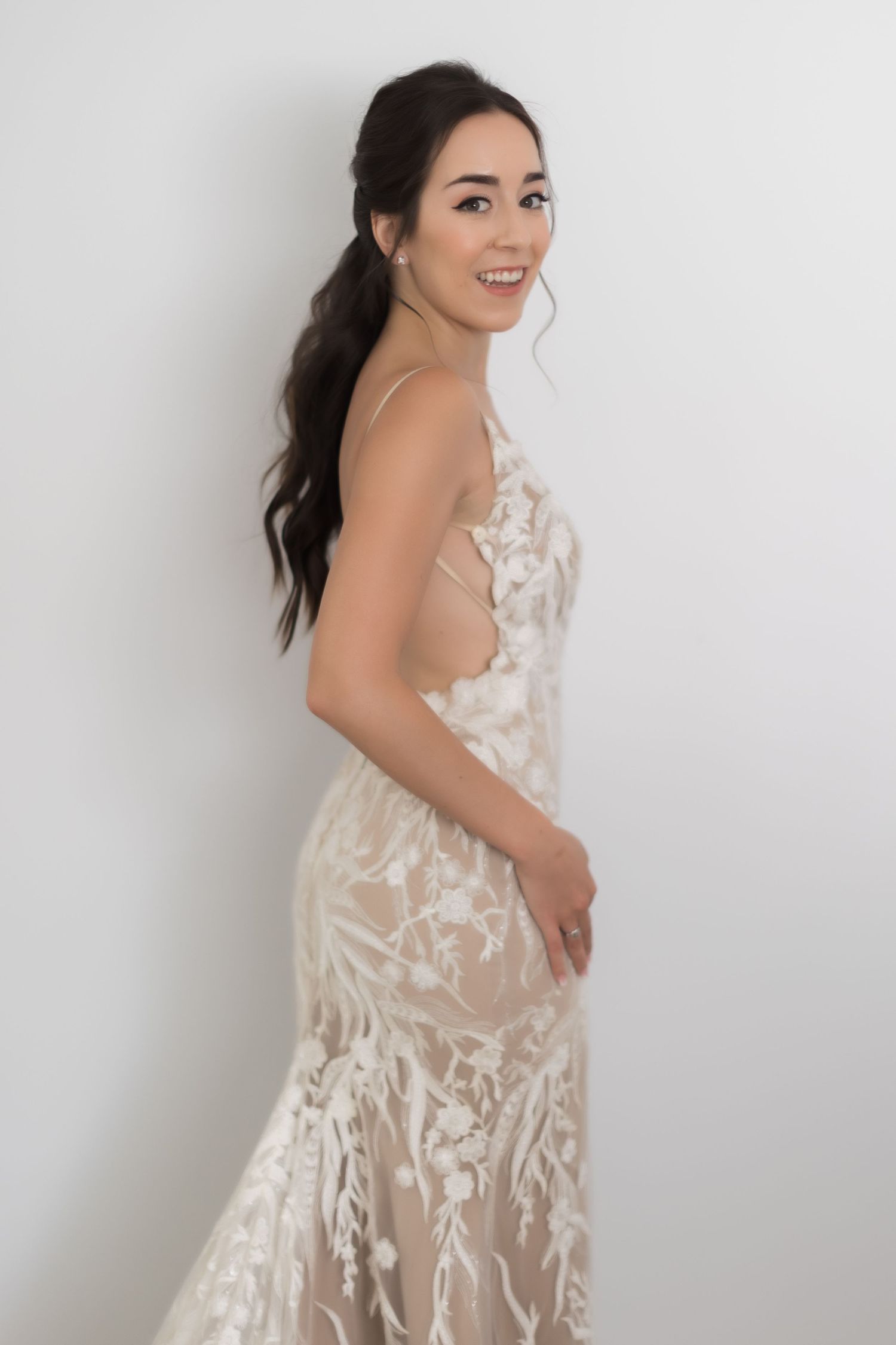Woman in white lace wedding gown poses elegantly against white backdrop.