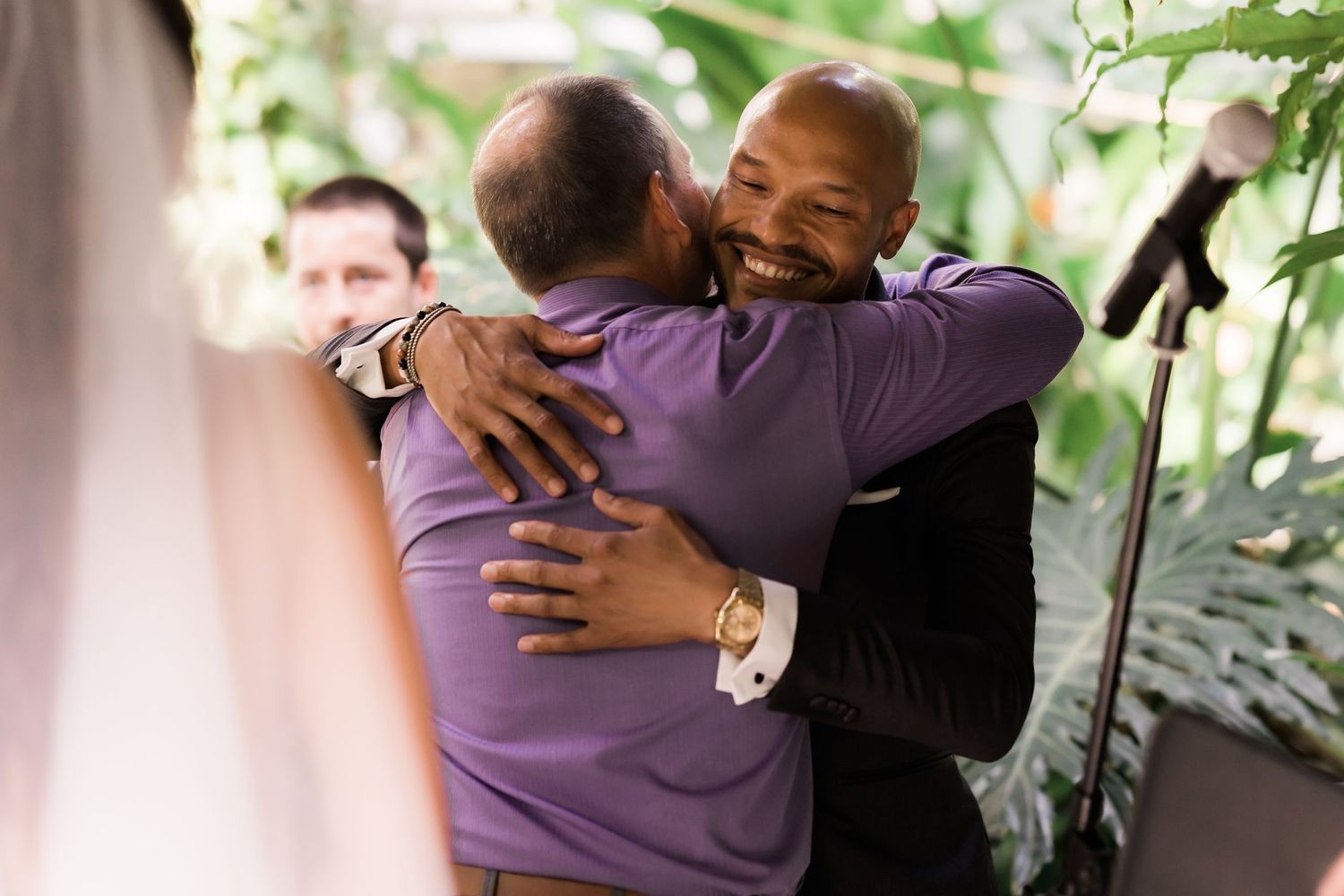Two people share an emotional hug during a wedding celebration surrounded by greenery.