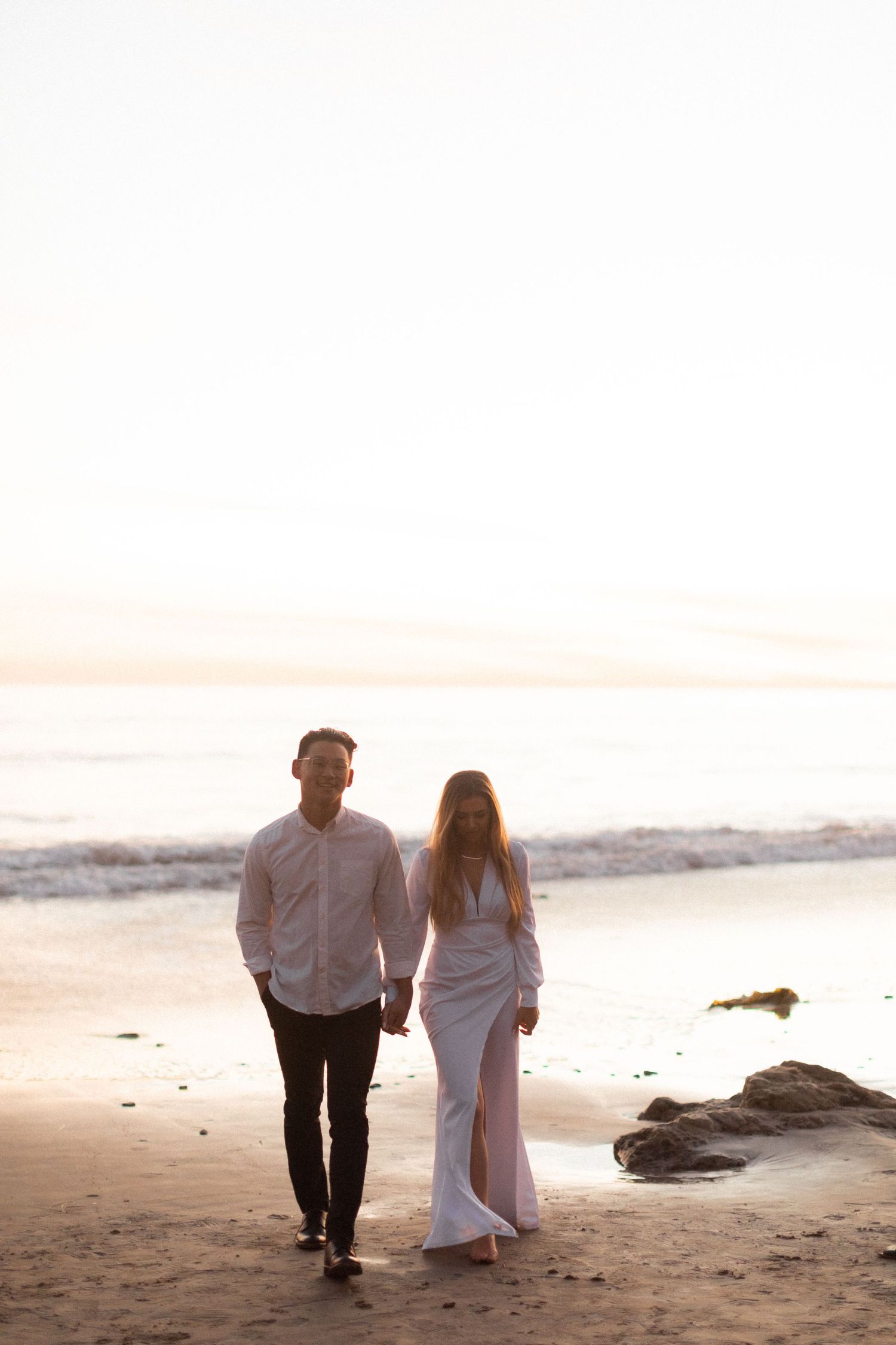 Couple walking hand in hand on sandy beach with ocean waves in background at dusk.