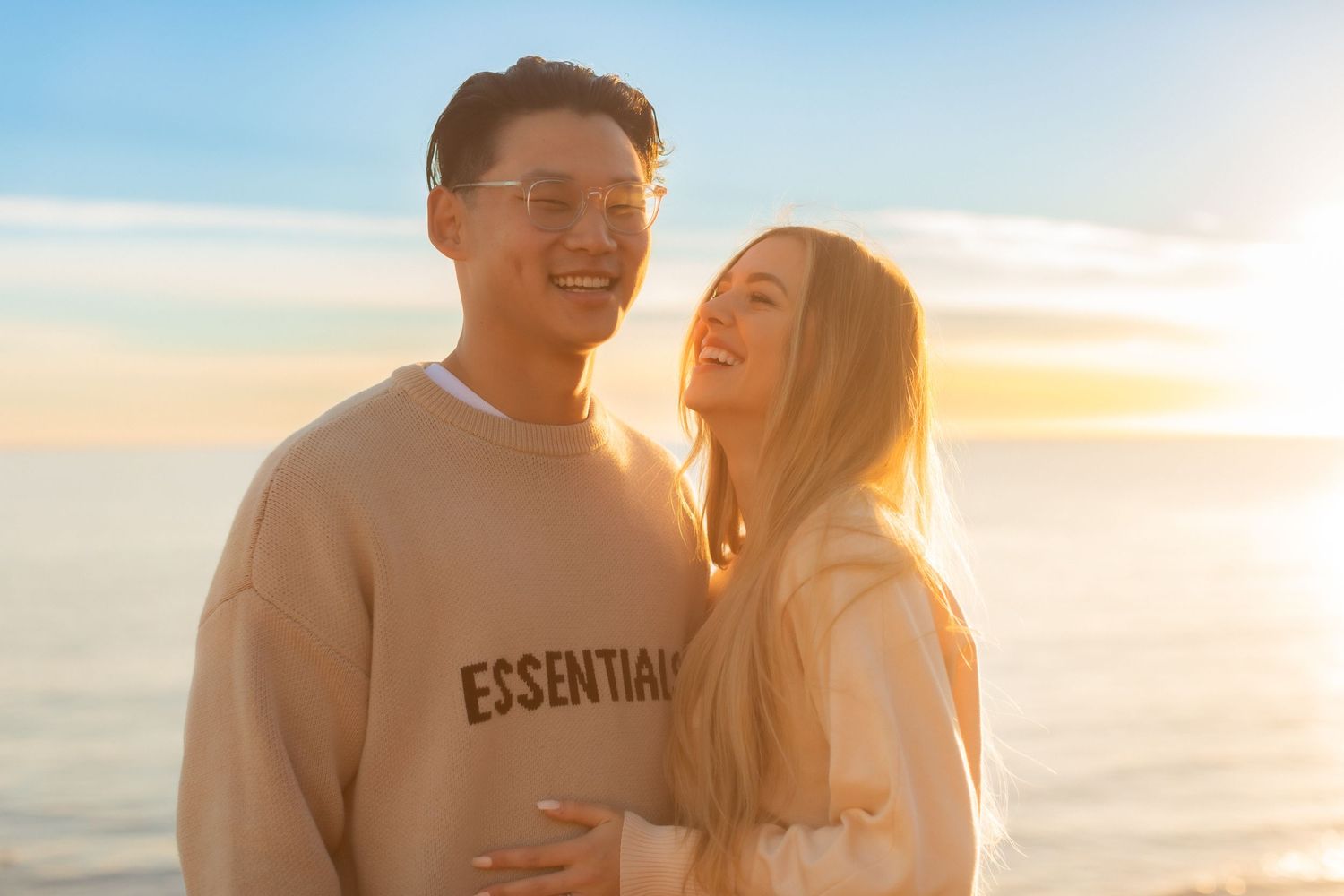 Couple shares a joyful moment at sunset by the ocean wearing matching light-colored outfits.