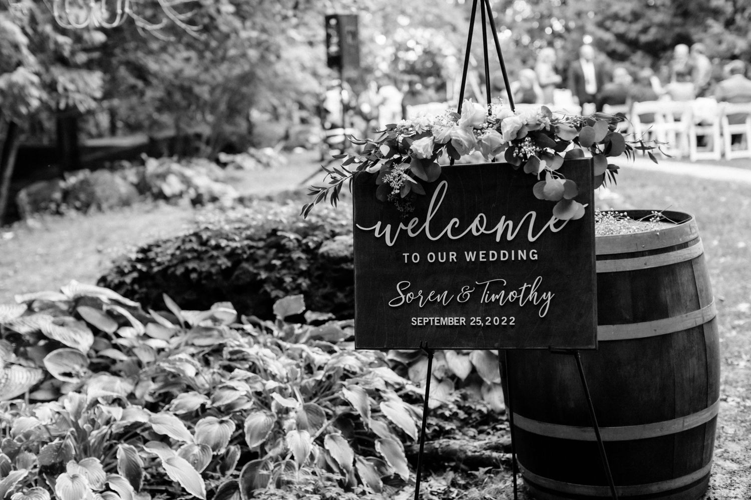 Black and white wedding welcome sign displayed on rustic barrel with floral decor.