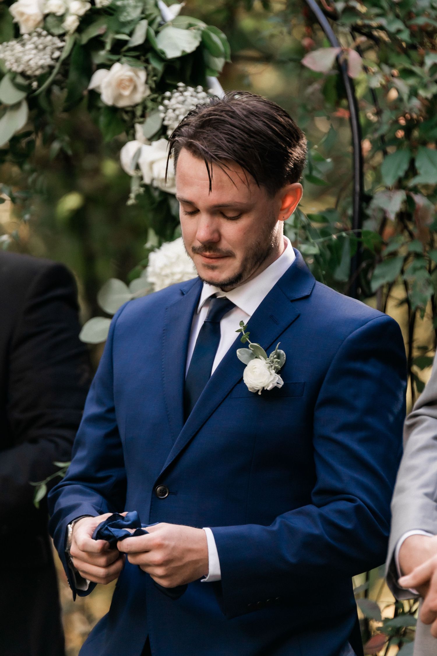 A groom in a navy suit holds a small object while standing under roses during a wedding ceremony.
