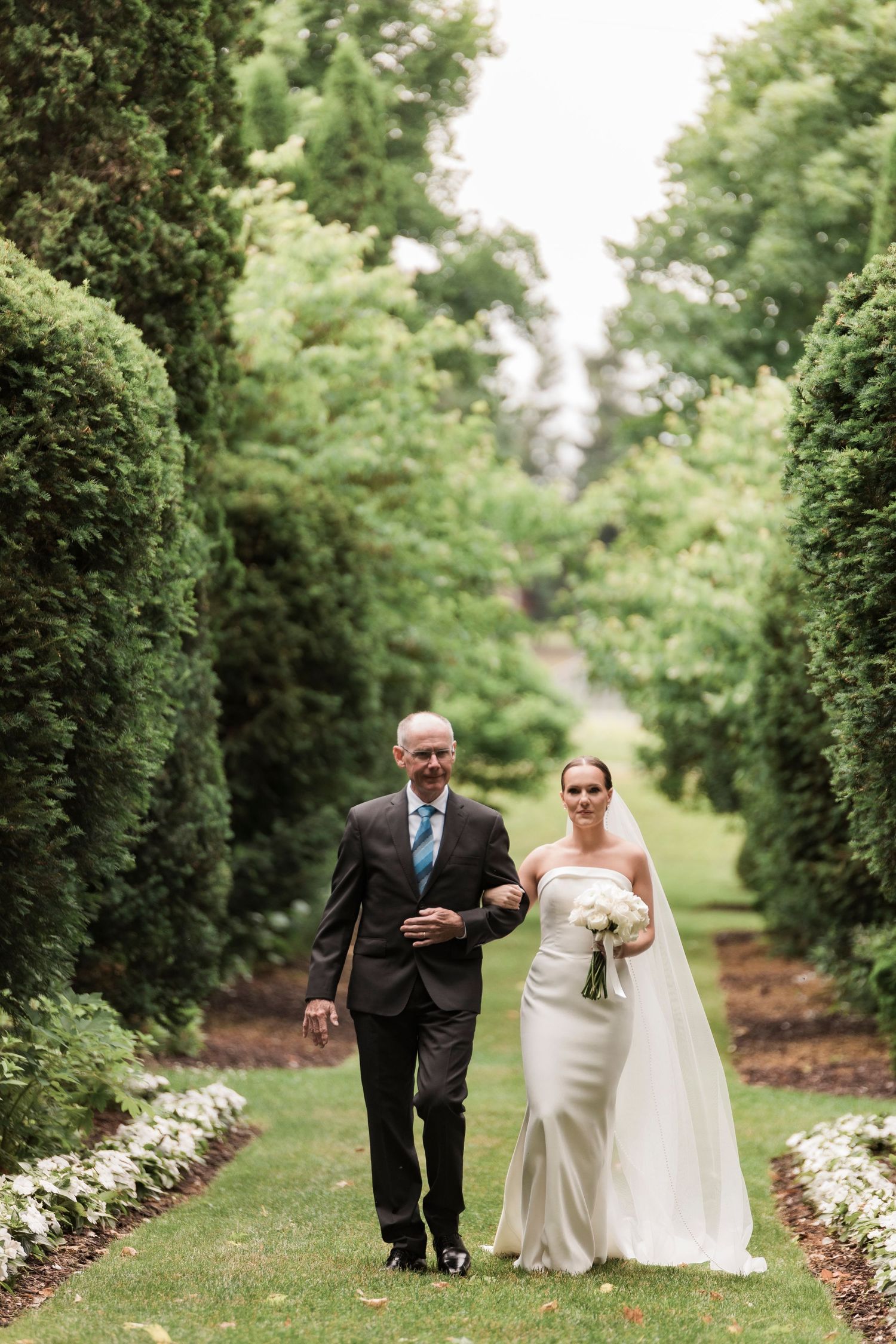 A couple in wedding attire walks along a scenic garden path lined with lush greenery and white flowering shrubs.
