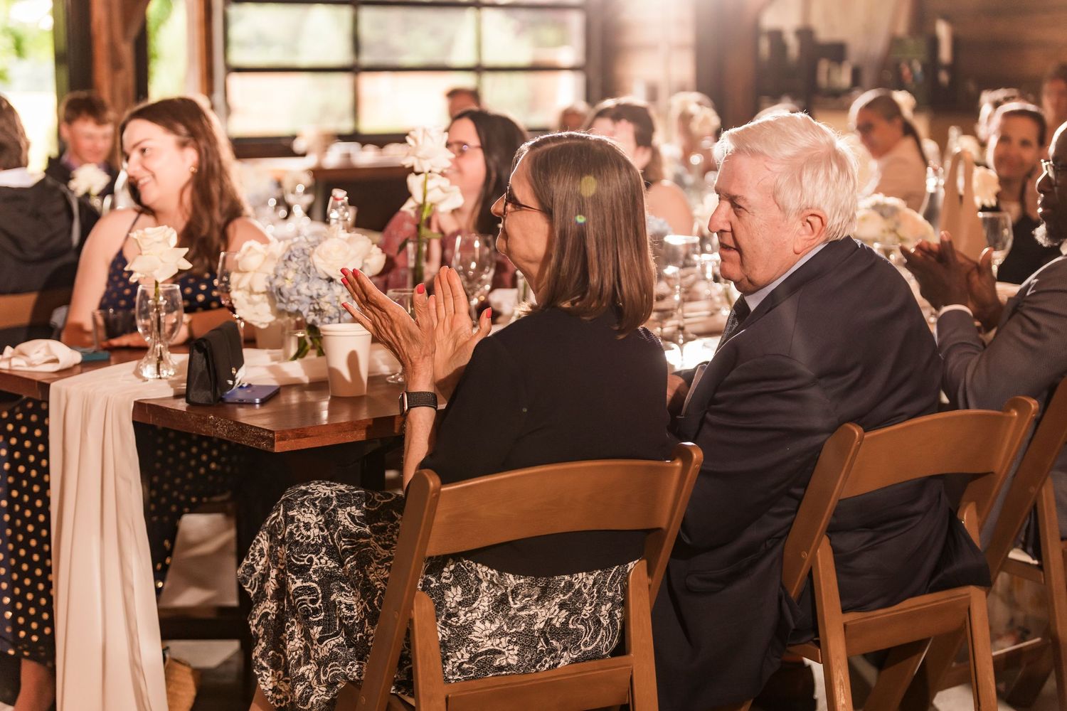 Wedding guests celebrate and applaud during a reception in a rustic wood-paneled venue with warm lighting.