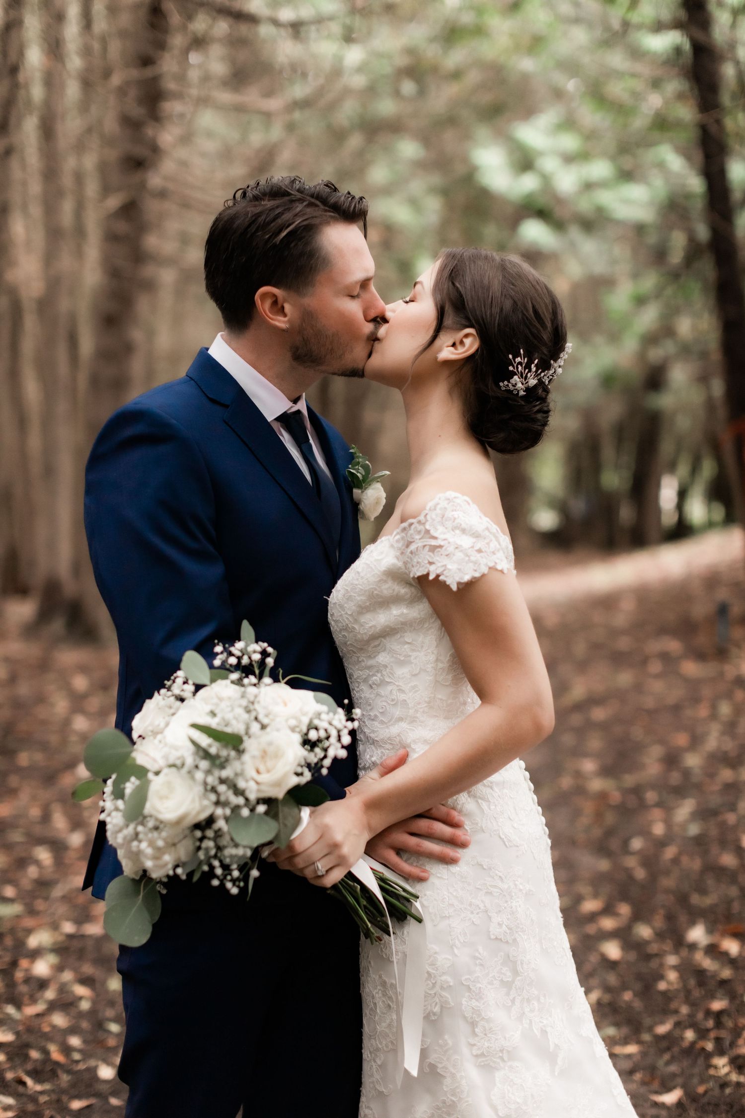 Newlywed couple sharing a romantic kiss in a wooded forest setting with white floral bouquet.
