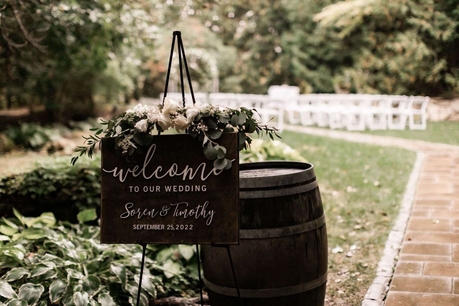 Rustic wooden barrel with welcome wedding sign decorated with greenery in an outdoor venue.