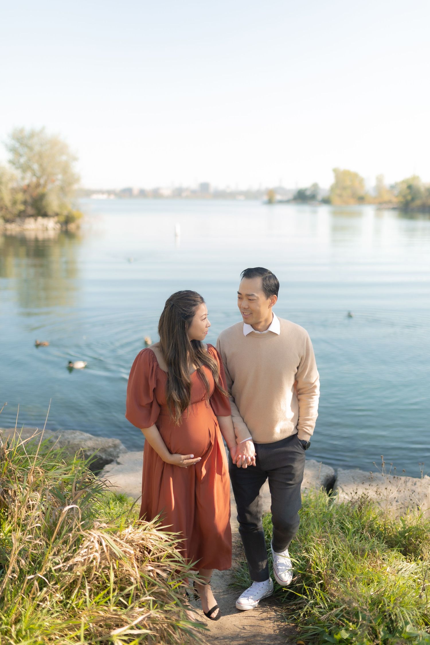 A couple holds hands while standing by a serene lake with ducks swimming in the background.