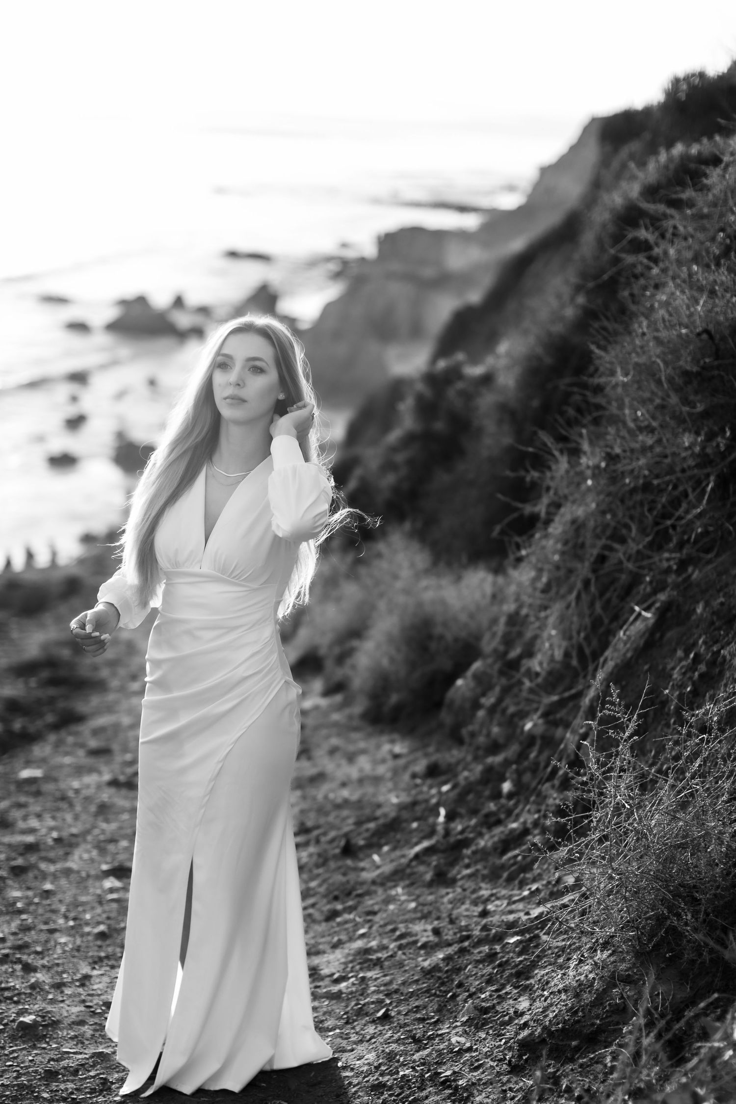 A woman in a white dress poses elegantly on a coastal cliff path during sunset.