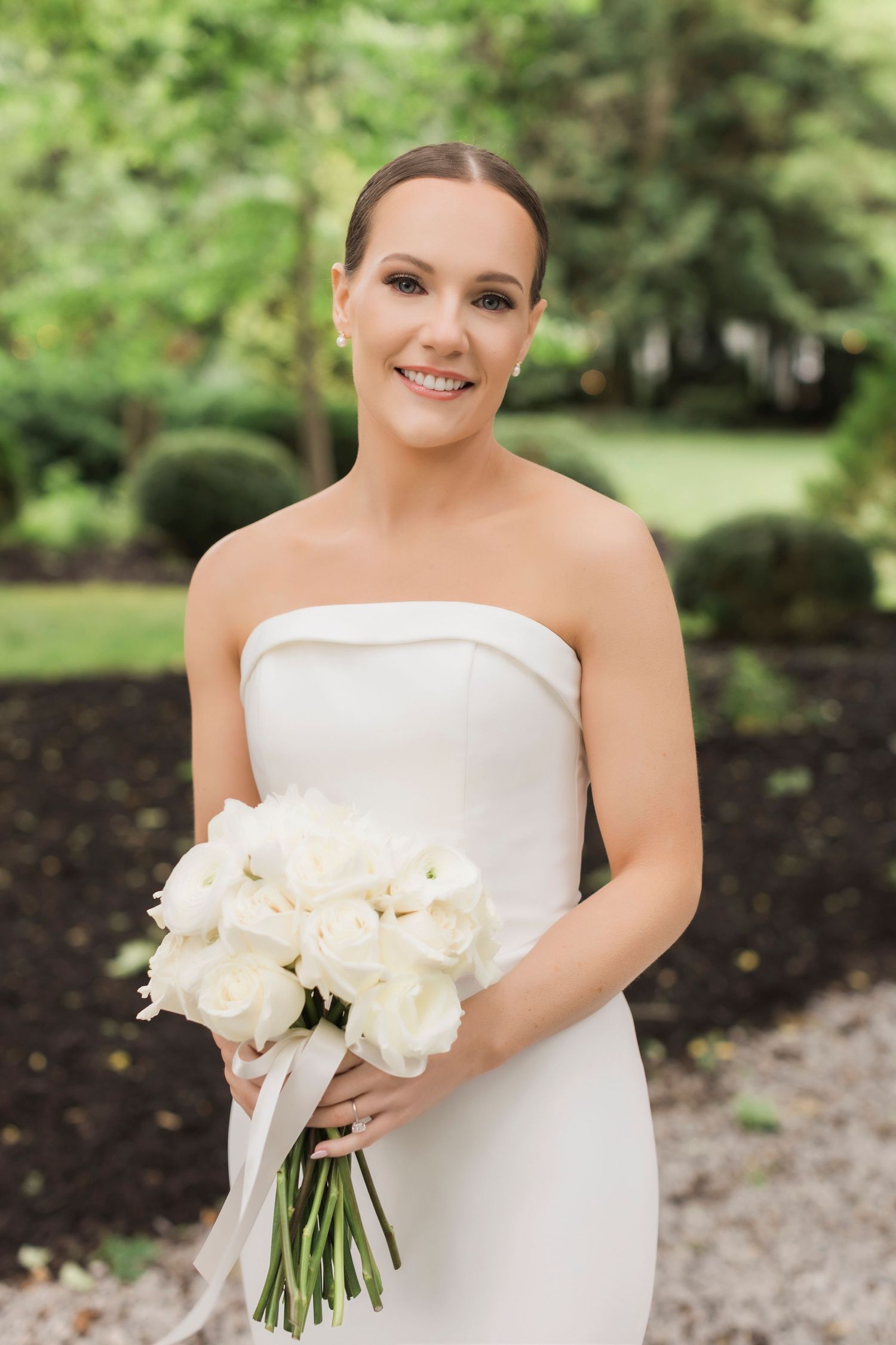Bride in minimalist strapless gown holds classic white rose bouquet in garden setting.
