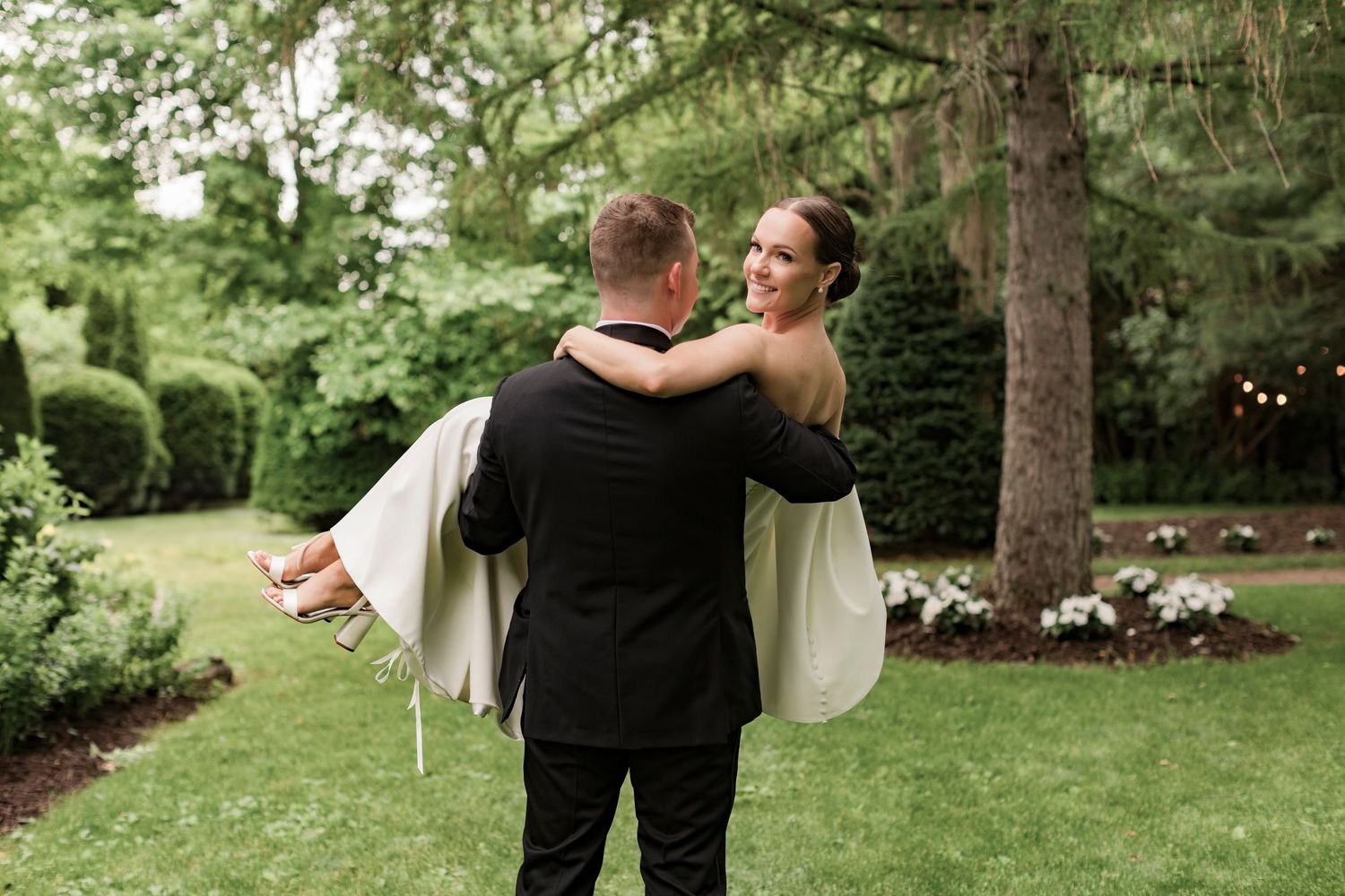 Joyful wedding moment captured as groom lifts bride in garden setting.