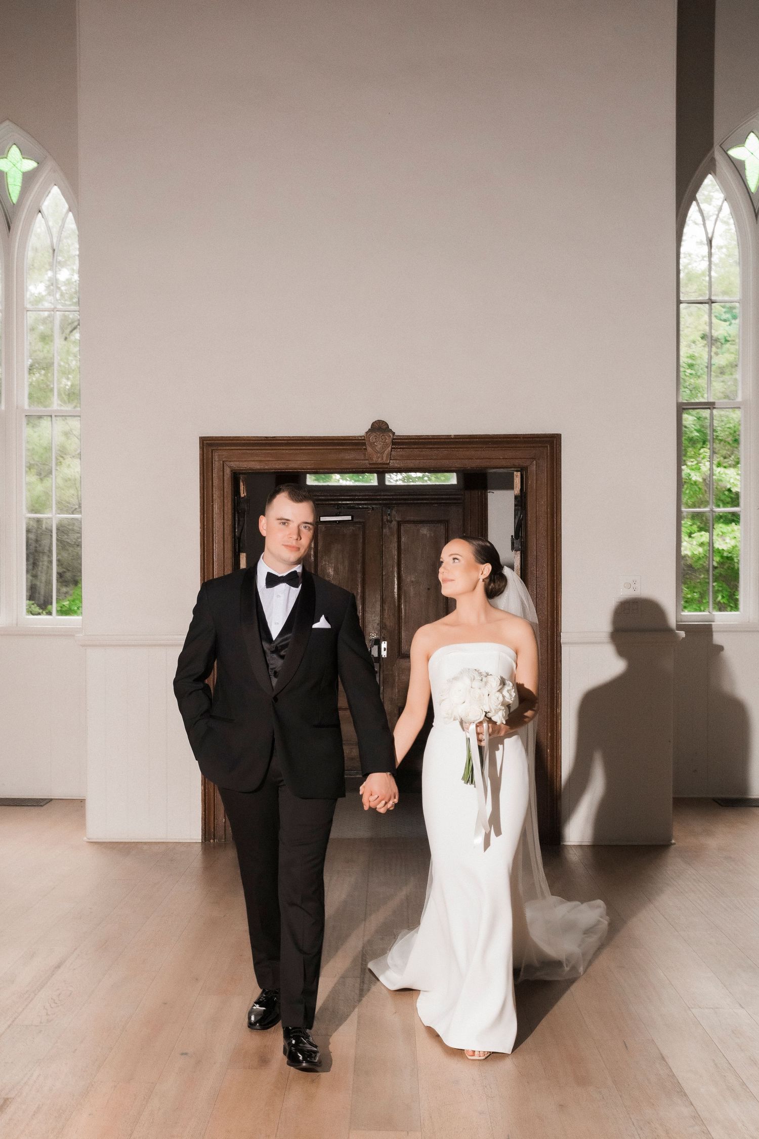 Newly married couple walking through an elegant church doorway with gothic windows and hardwood floors.