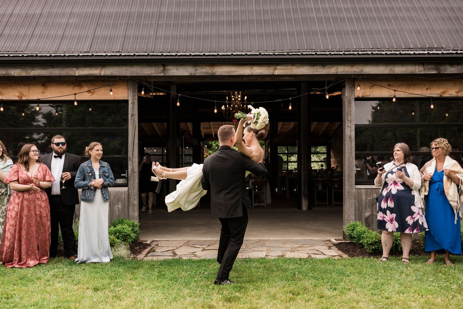 A couple embraces in front of a rustic barn entrance while wedding guests look on from both sides of the doorway.