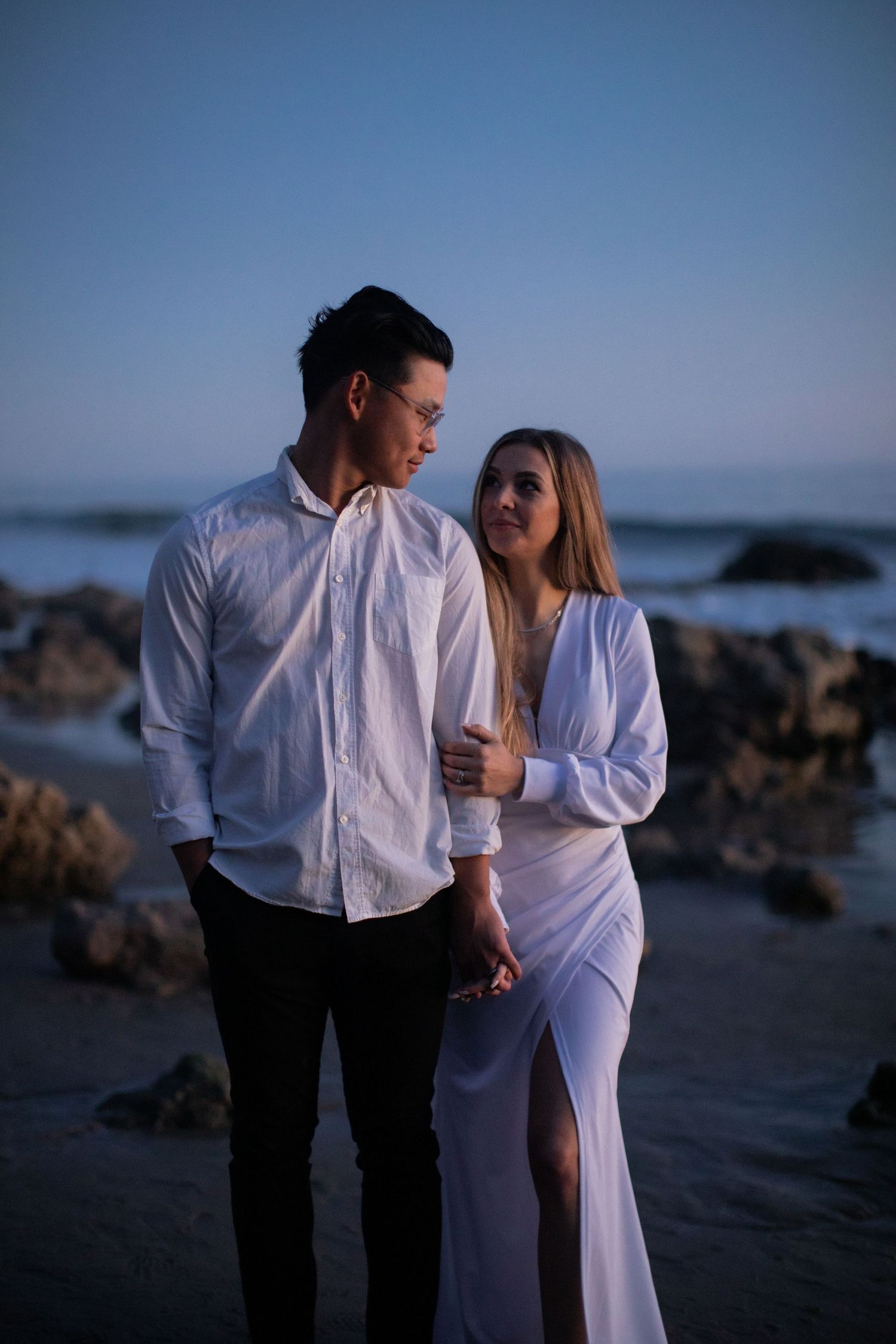 Coastal engagement photo of couple in matching white outfits against ocean backdrop.