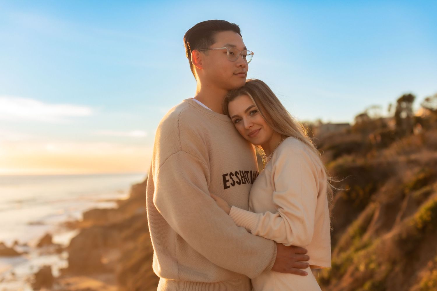 Romantic couple in matching cream sweaters share intimate moment on coastal cliff at golden hour.