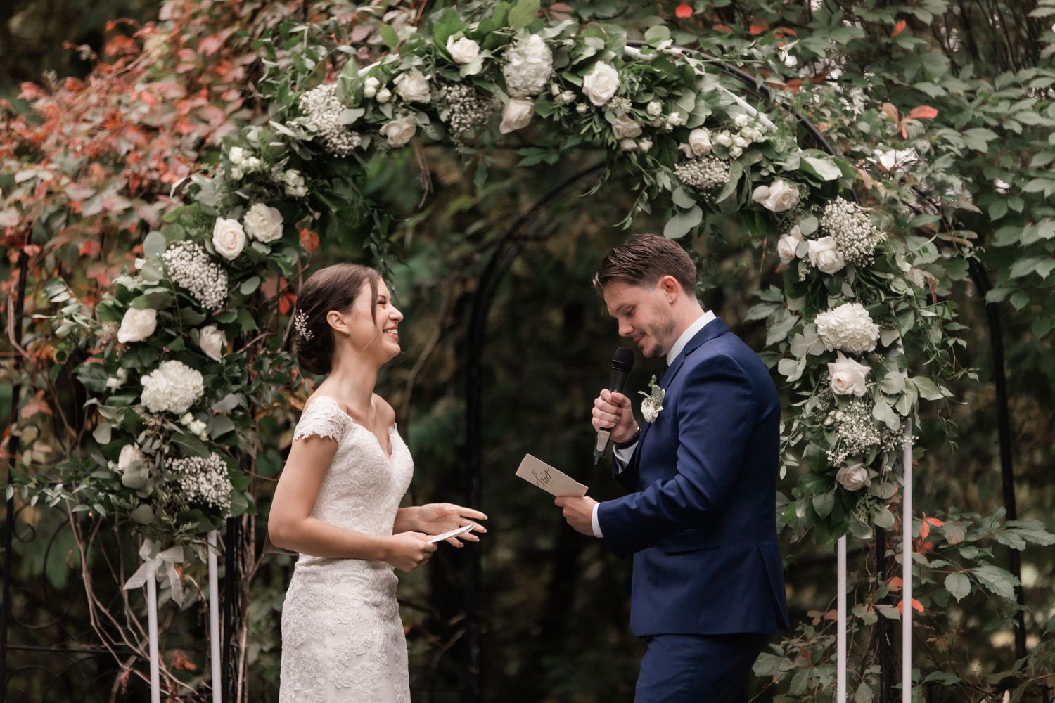 A couple shares an emotional moment while reading vows under a flower-covered arch.