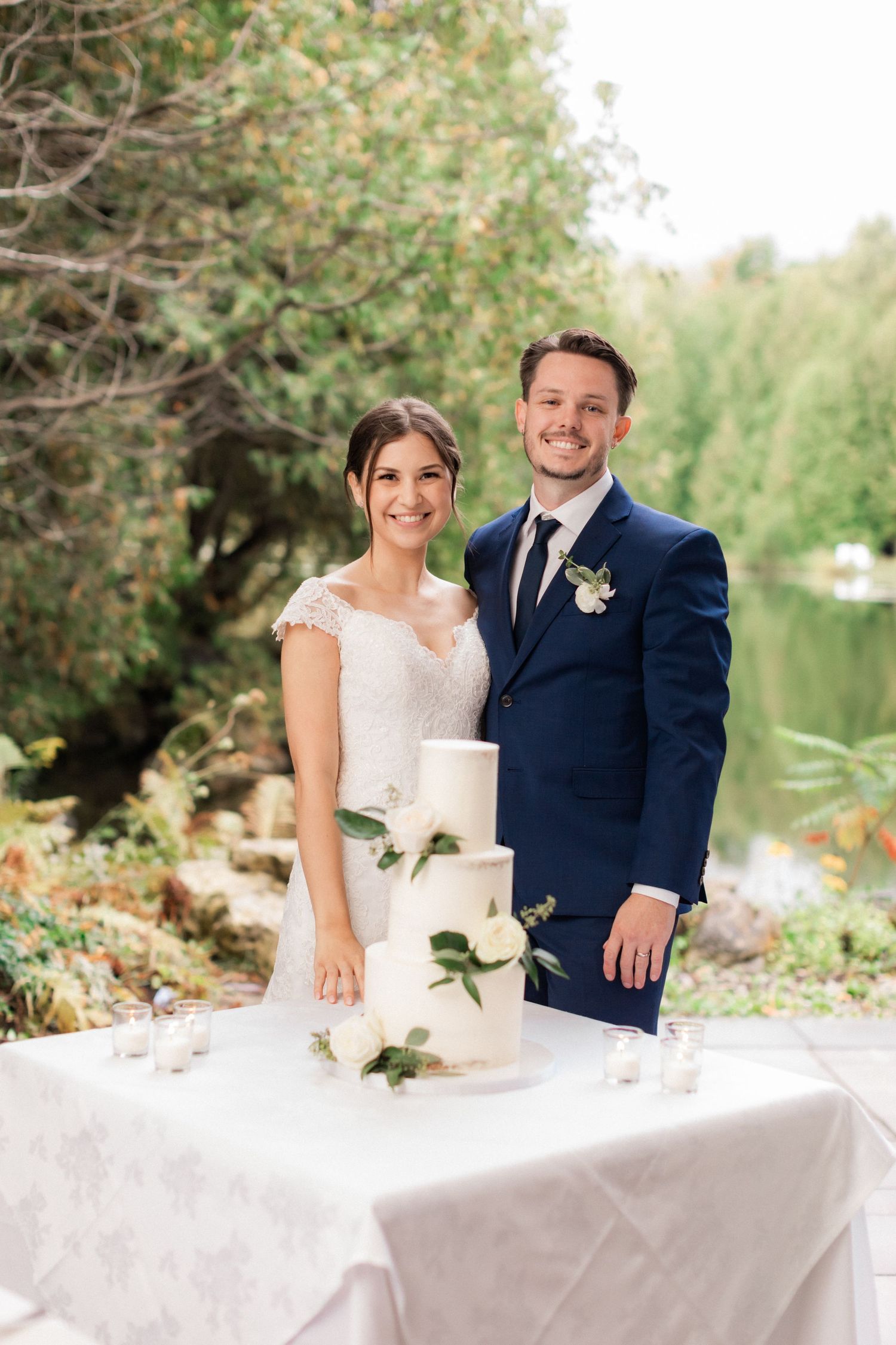 Wedding couple poses with elegant three-tier white cake decorated with roses.