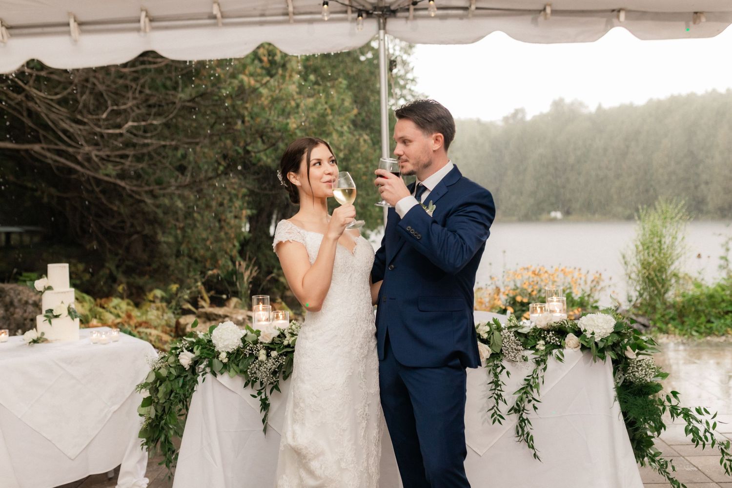A wedding couple shares a candid moment under a decorated tent by a lake.