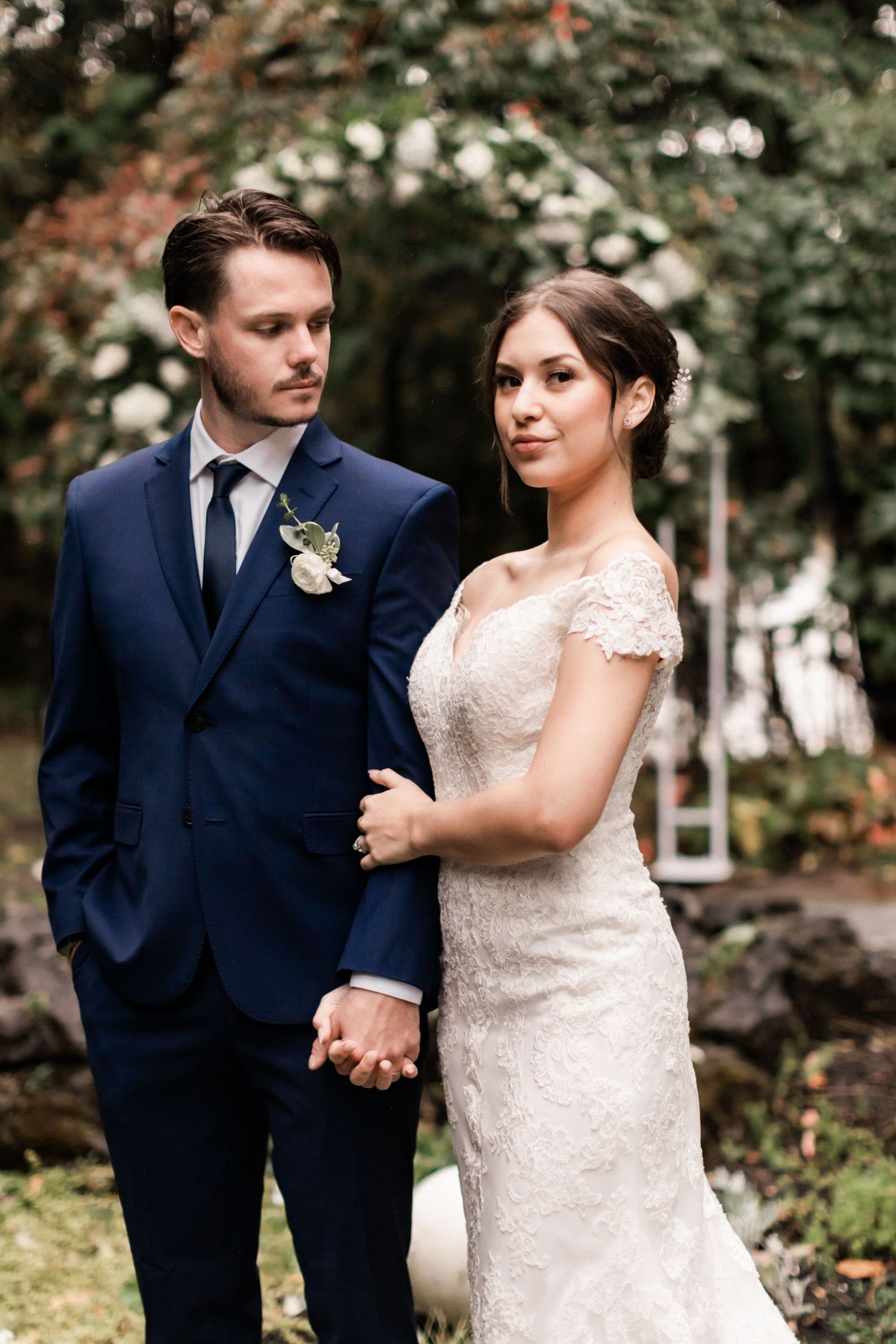 Elegant wedding portrait with couple in navy suit and lace dress against garden backdrop.