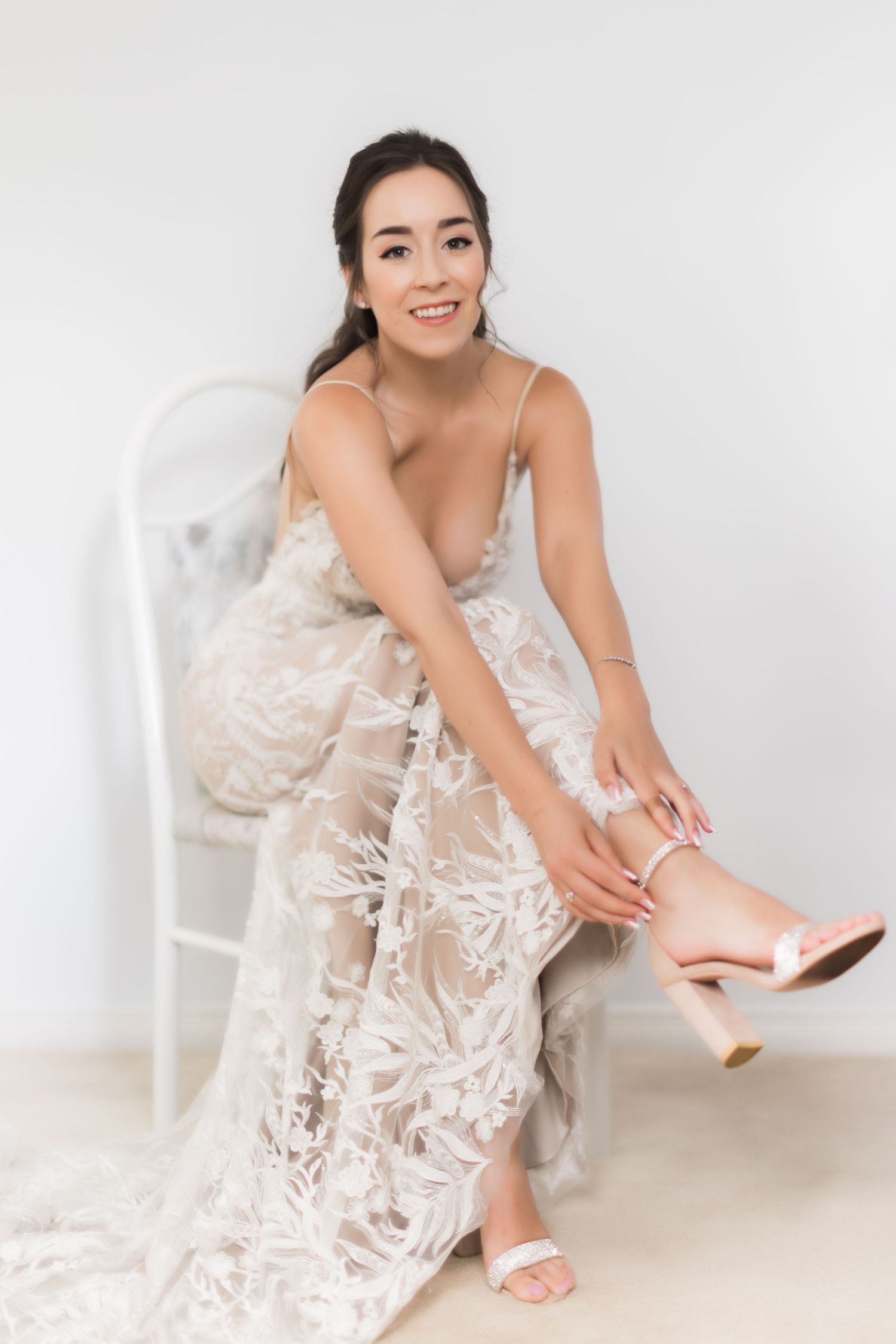 Bride in sheer lace wedding dress adjusts her shoe while sitting on white chair.