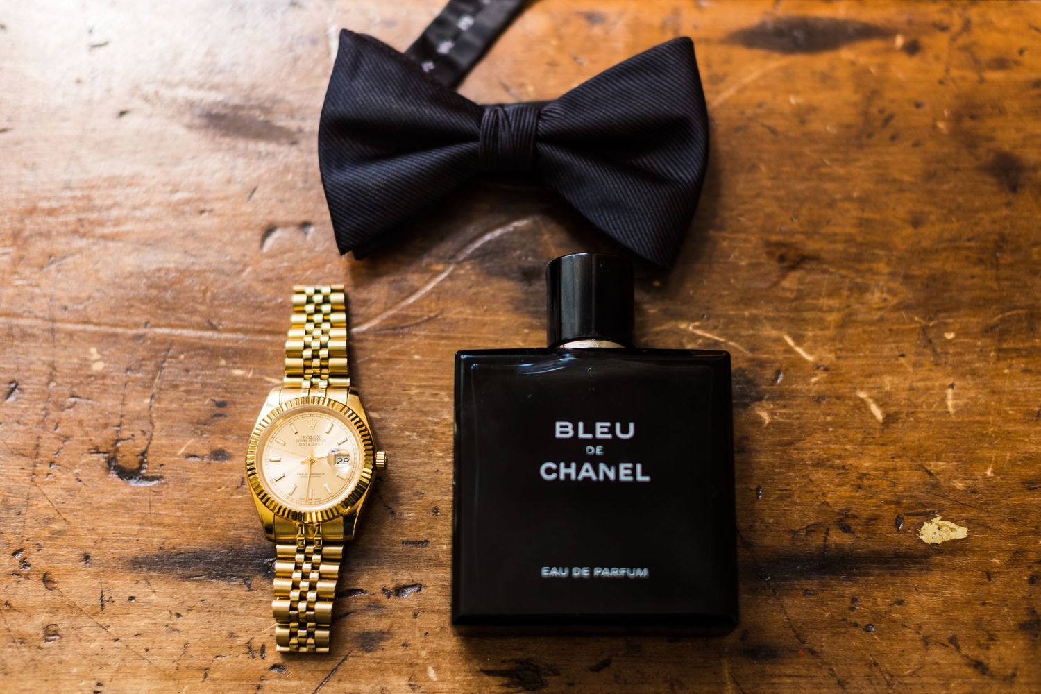 Black bowtie, gold watch, and Chanel Bleu cologne bottle arranged on wooden surface.