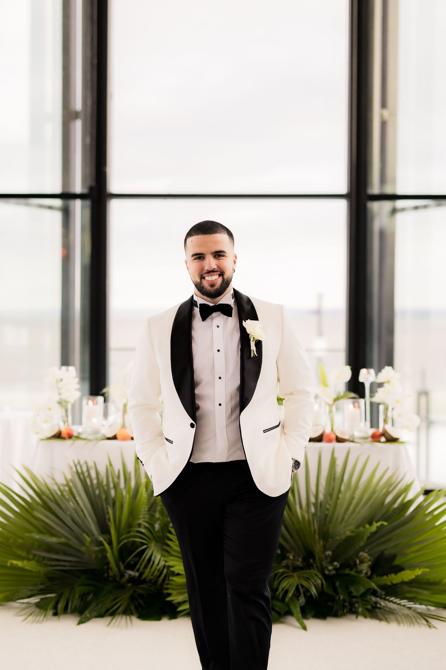 Groom in white dinner jacket and black bow tie poses in front of window and tropical plants.