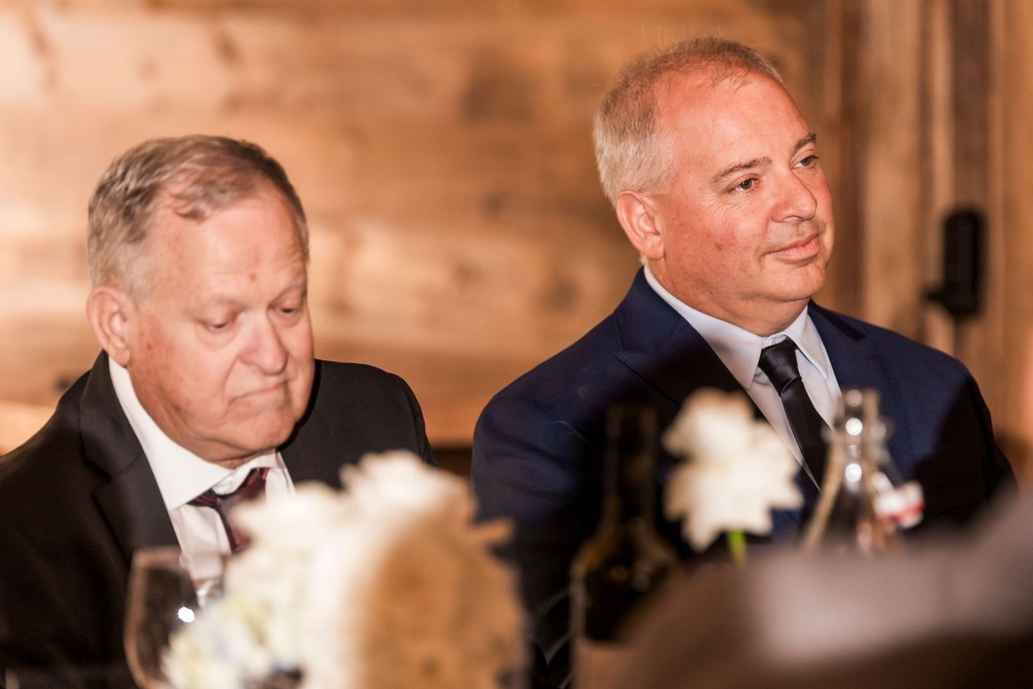 Two men in dark suits sit at a formal event with white floral decorations in the foreground.