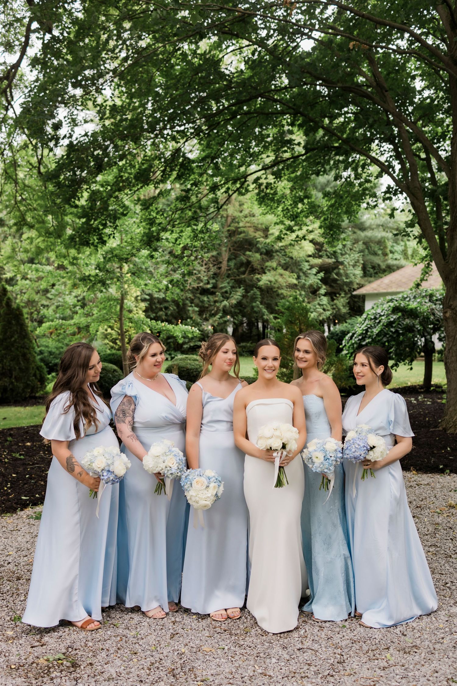 A bridal party in light blue bridesmaid dresses and a white wedding gown pose together on a gravel path under green trees.