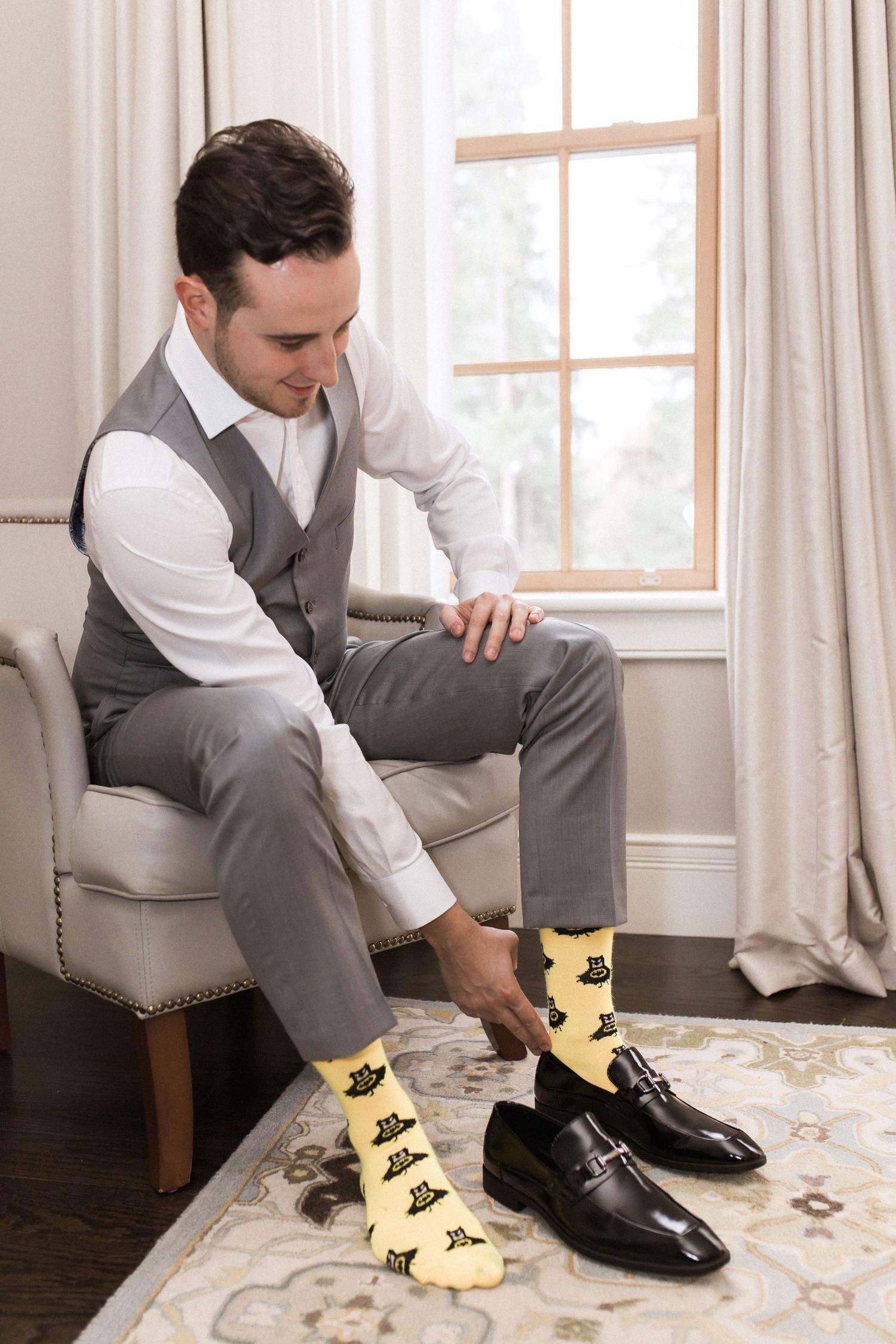 Groom in grey suit and vest puts on fun patterned socks and dress shoes while getting ready.
