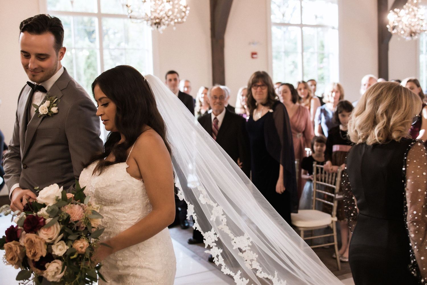 Bride in white dress with long veil standing next to guest in sparkly dress during ceremony.