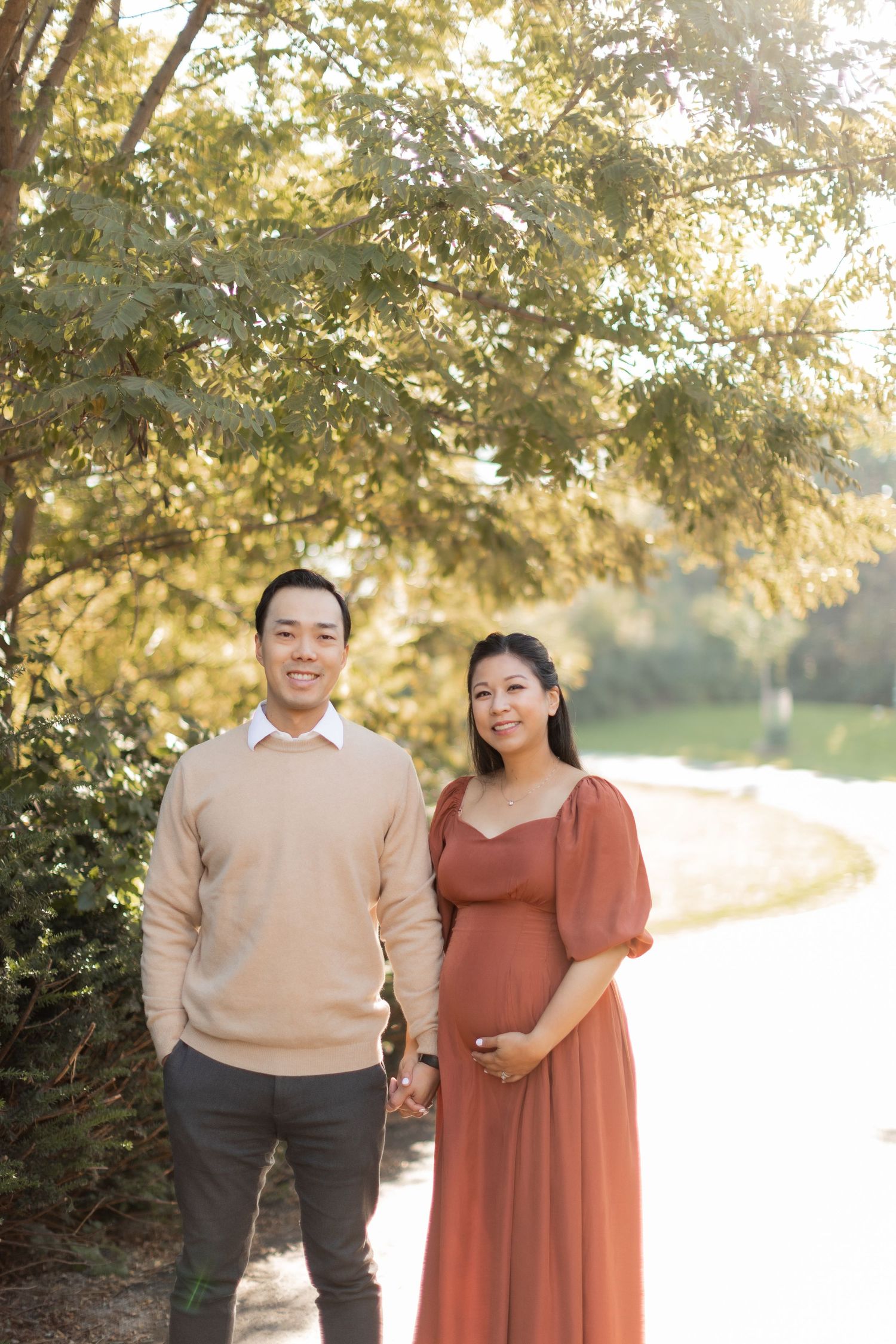 A sunny outdoor maternity portrait with a couple in coordinated casual wear on a path.