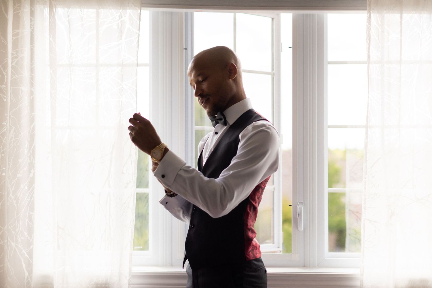 A silhouetted figure in formal attire adjusts clothing by a bright window.
