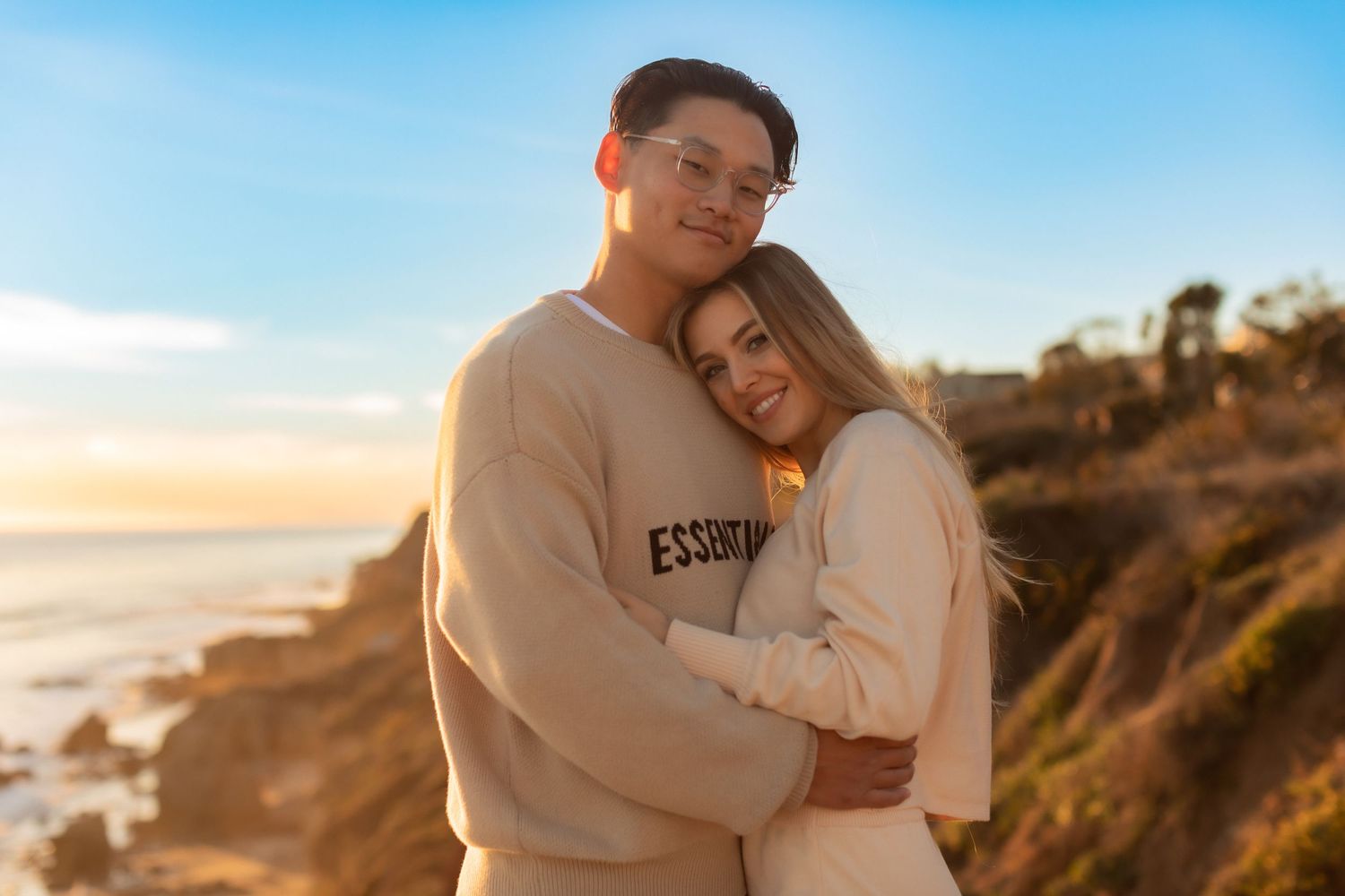 Couple in casual cream sweaters hugging and smiling on scenic coastal overlook.