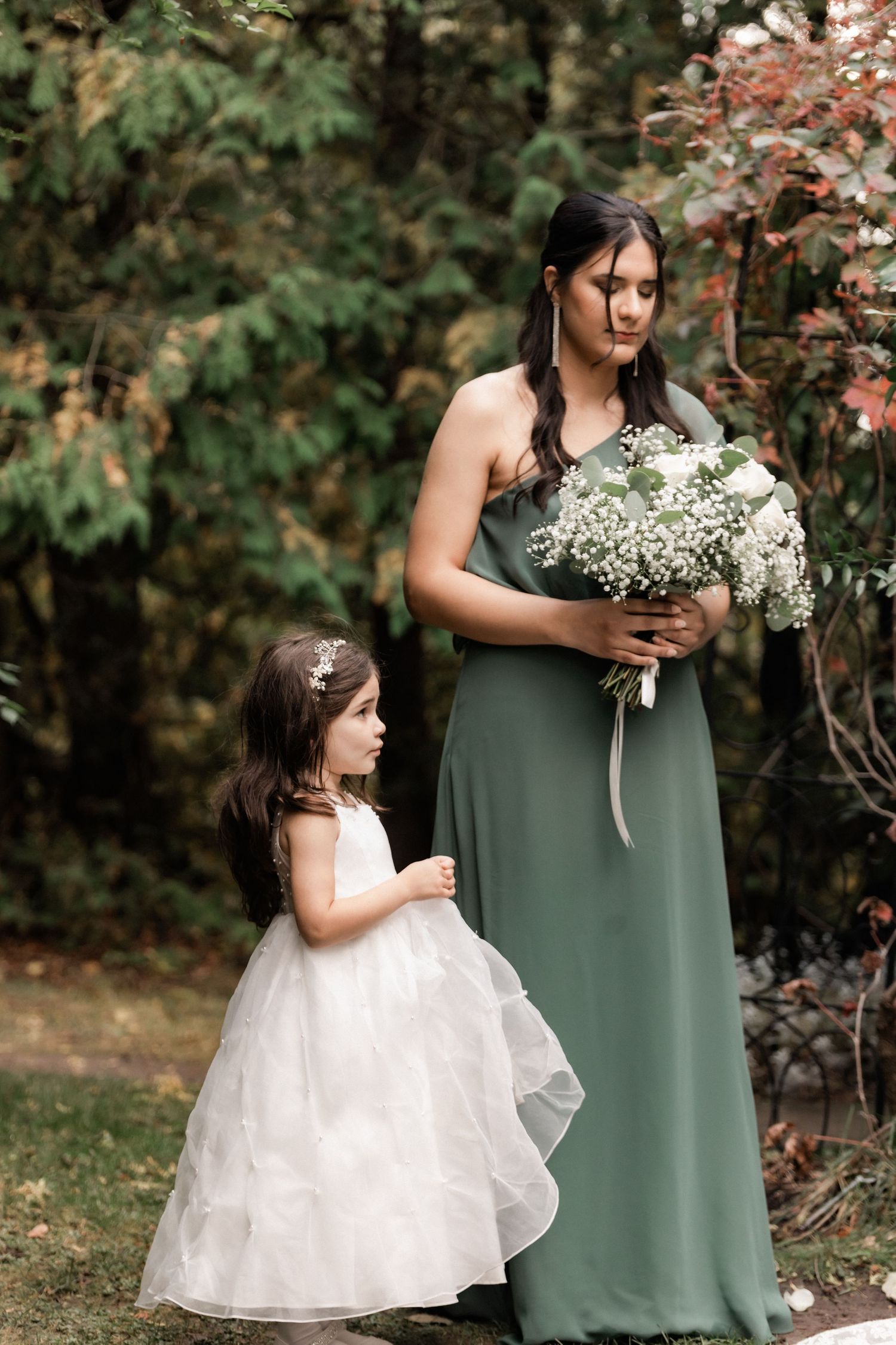 A bridesmaid in sage green dress holds baby's breath bouquet while standing with flower girl in white dress.
