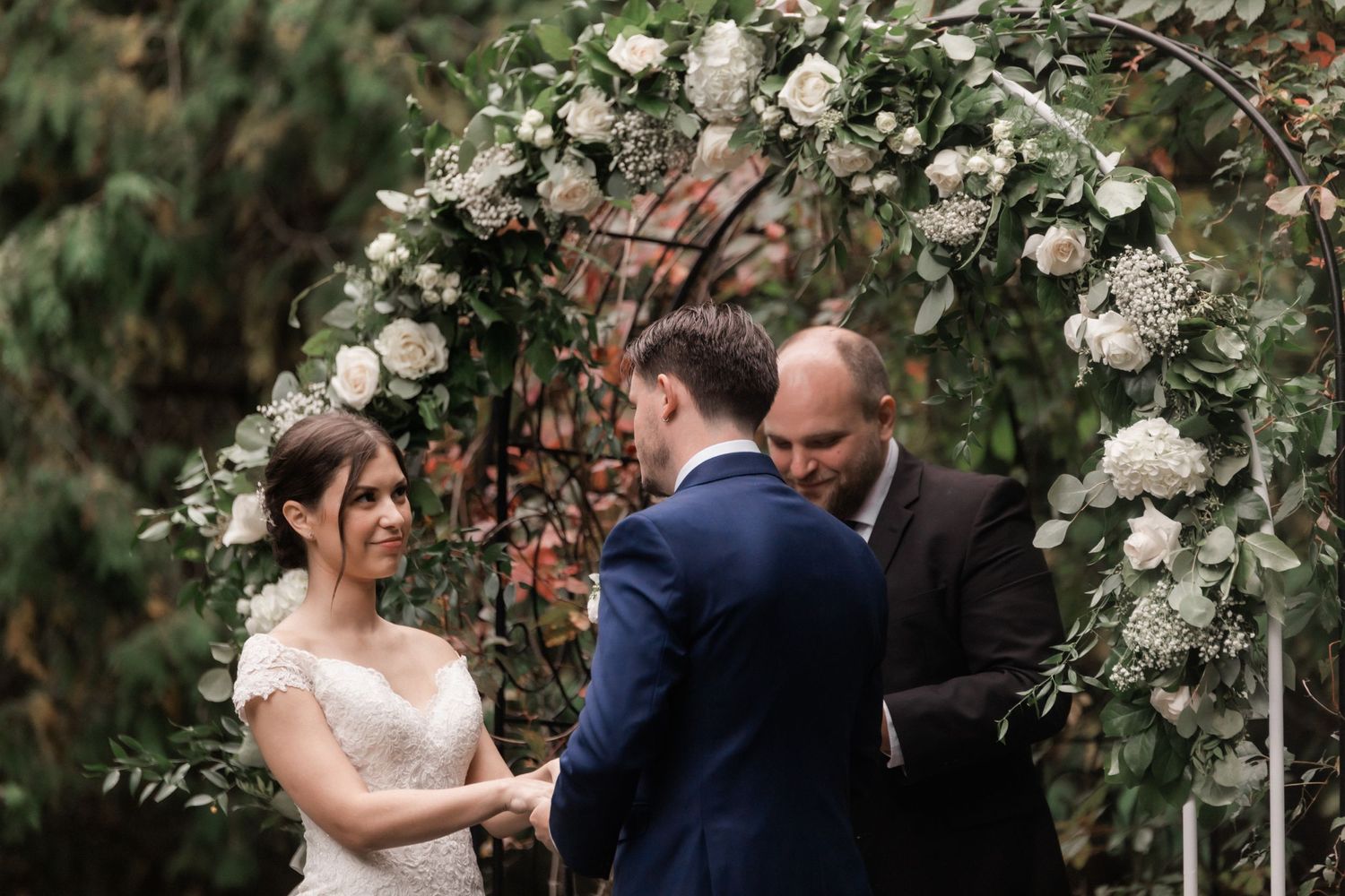 A couple exchanges vows under a white floral arch during their outdoor wedding ceremony.