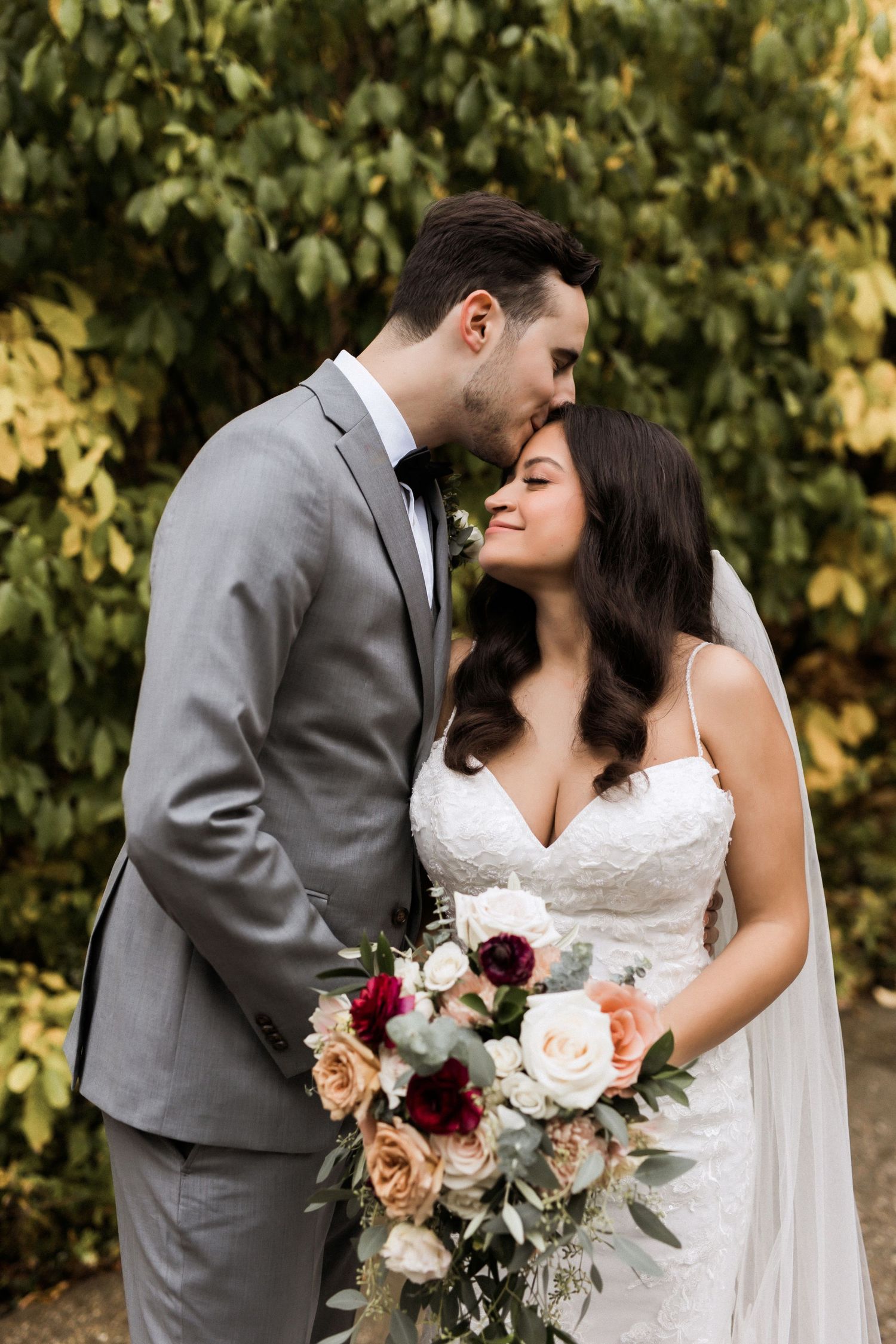 Wedding couple shares tender moment with elegant bouquet of burgundy and blush roses.