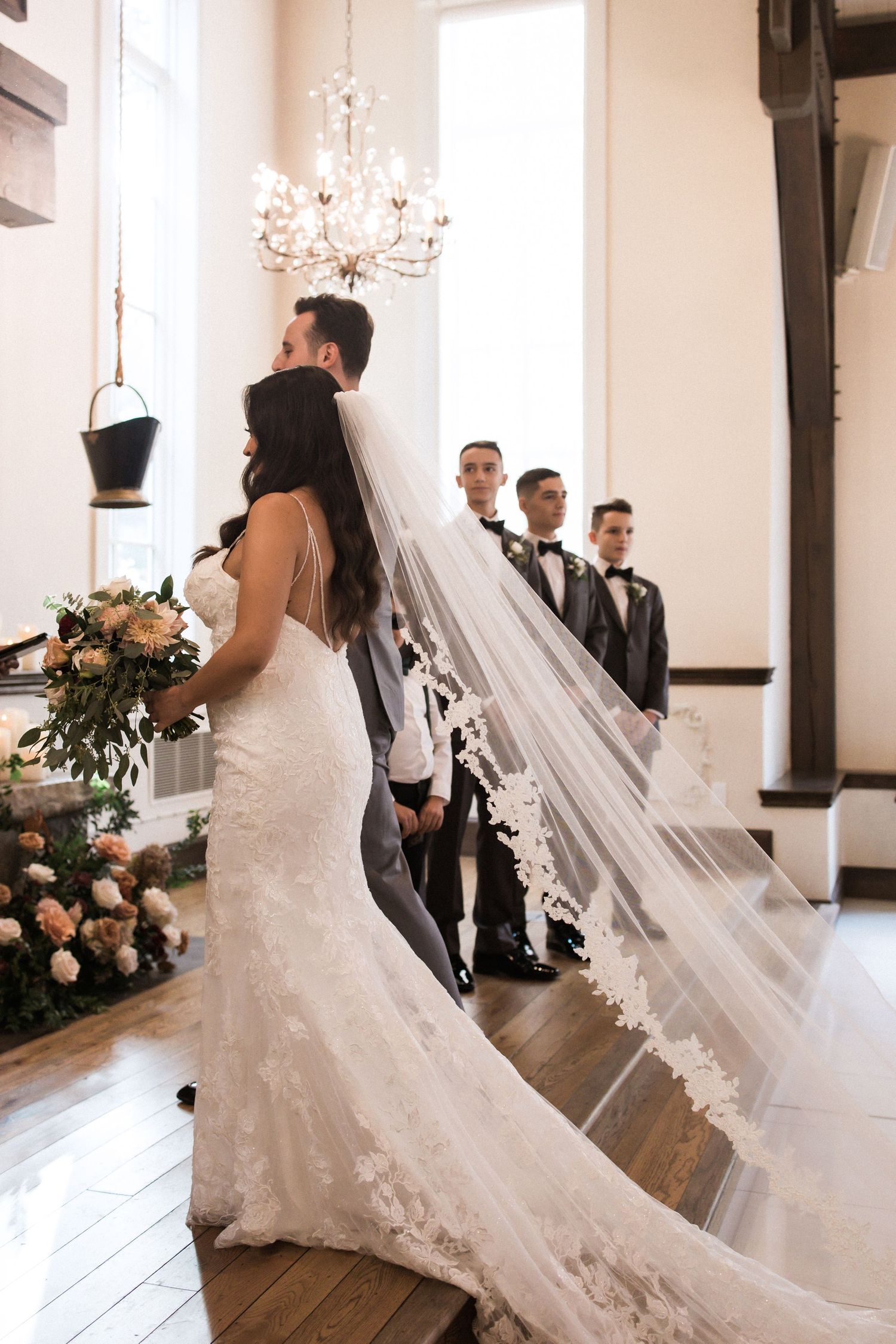 Bride in fitted lace gown with flowing veil stands with groomsmen in formal attire.