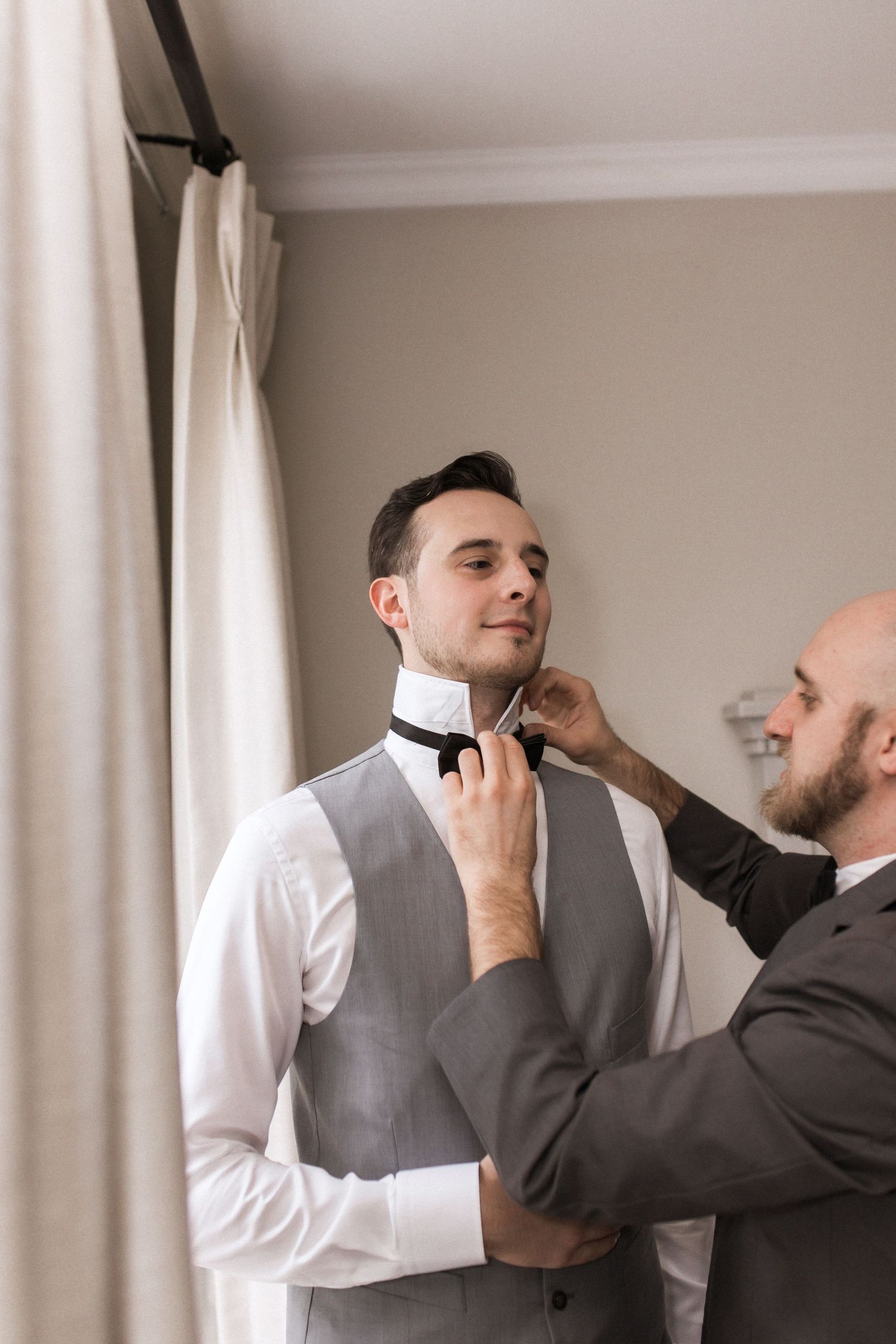 Helper adjusts groom's bowtie as final wedding preparation touch.