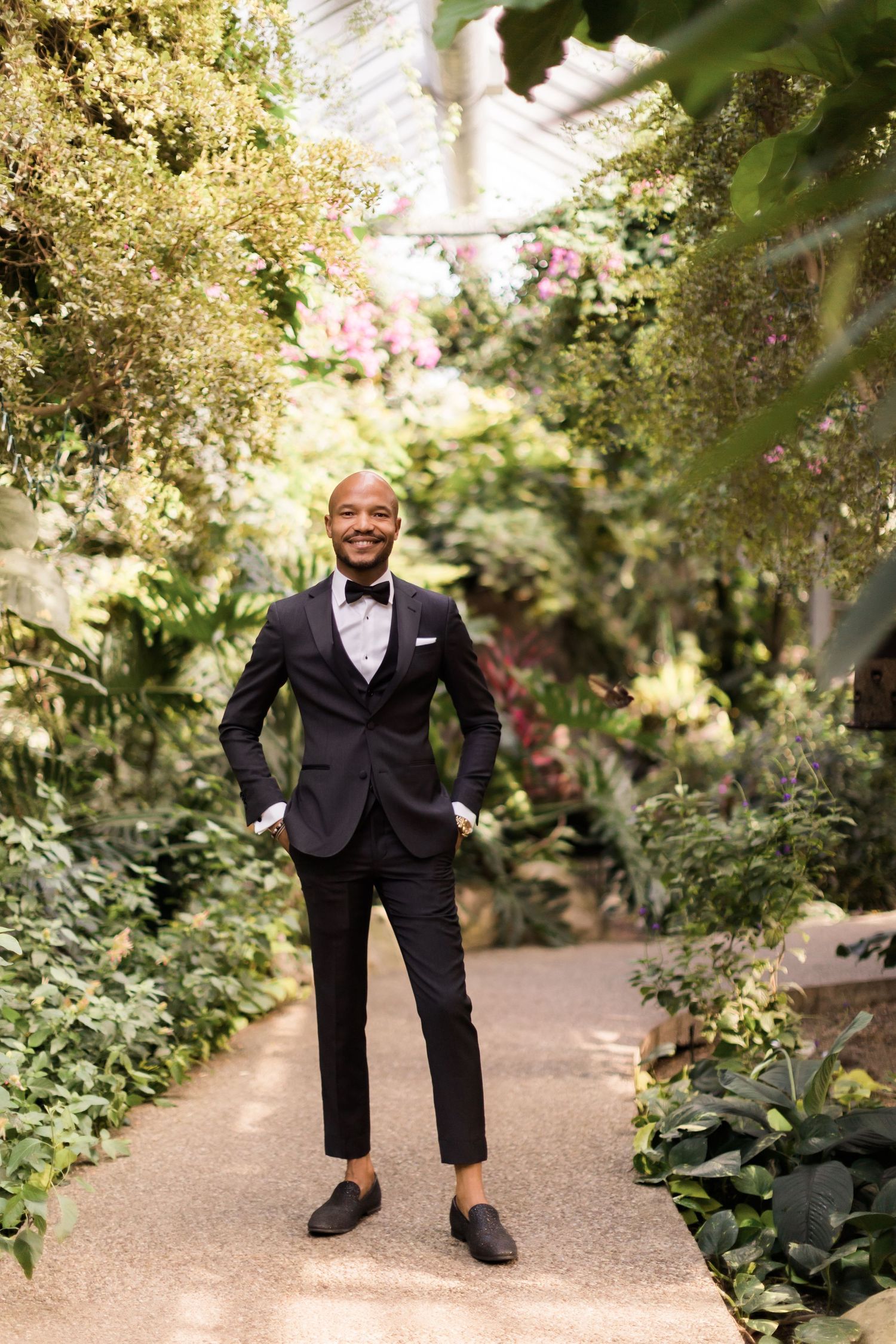 Elegantly dressed person in a navy suit stands on garden path surrounded by lush greenery.