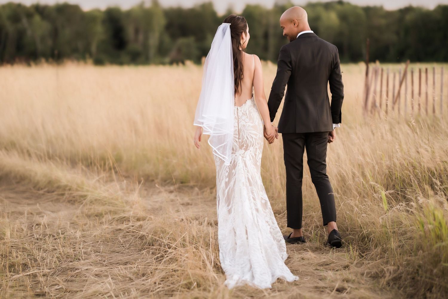 Bride and groom walking away into golden field during sunset, showing backs of wedding attire.