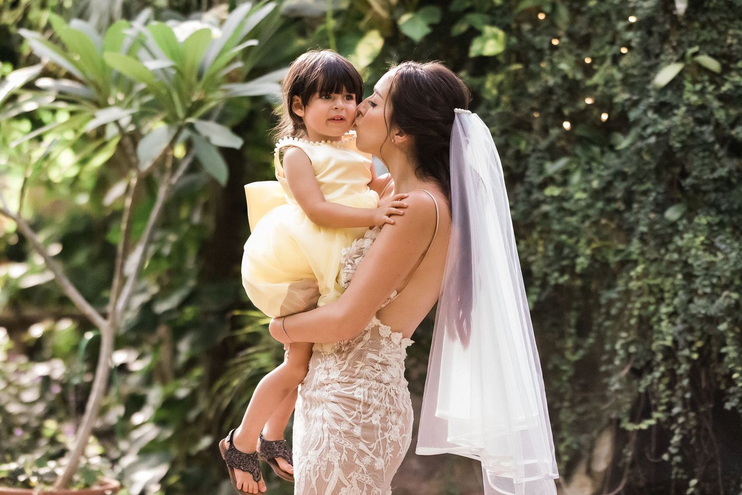 Bride in lace wedding dress holding young child in yellow dress during outdoor celebration.